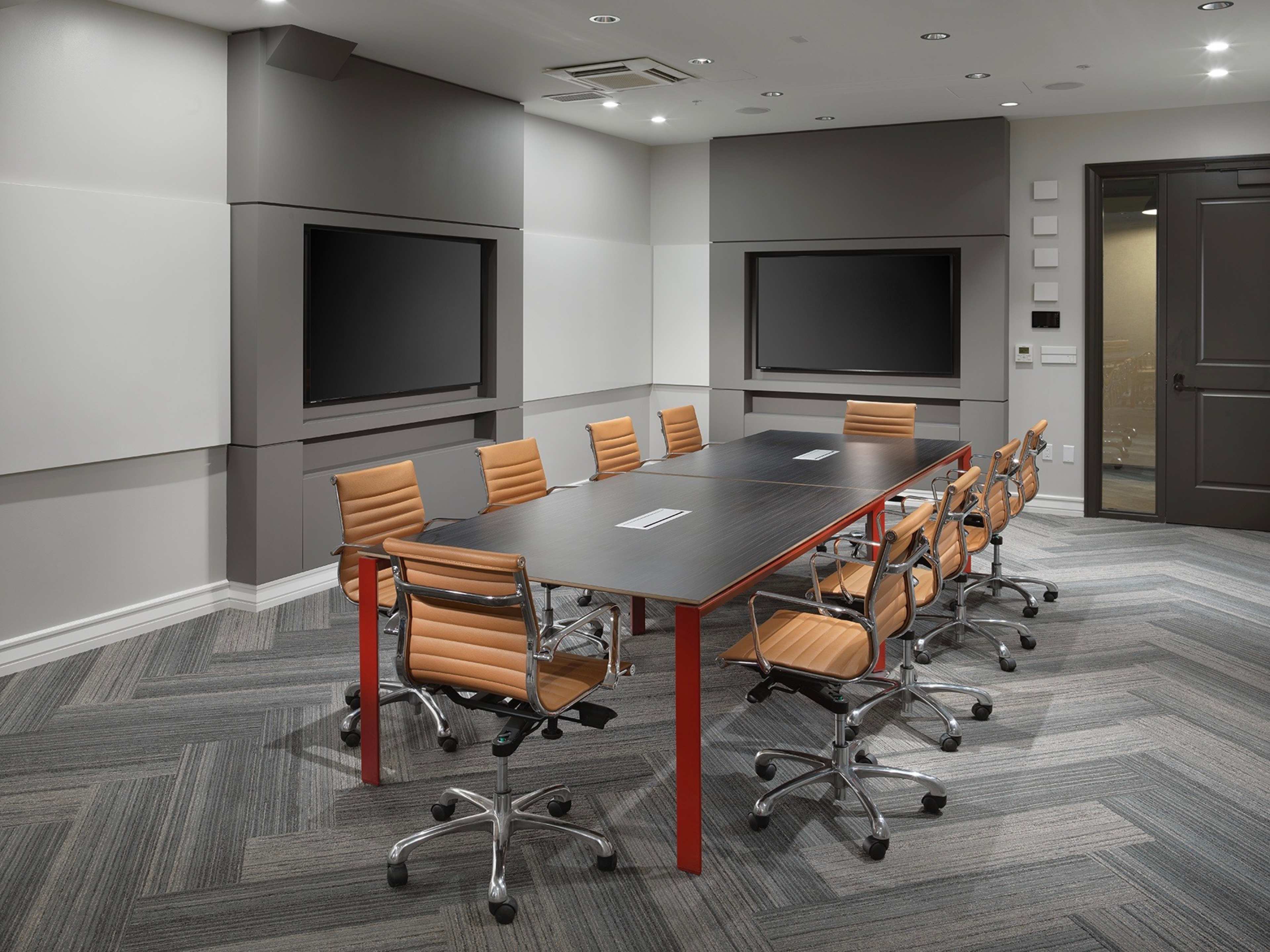 A modern conference room features a large table surrounded by orange office chairs, with two wall-mounted screens and soundproofing panels.
