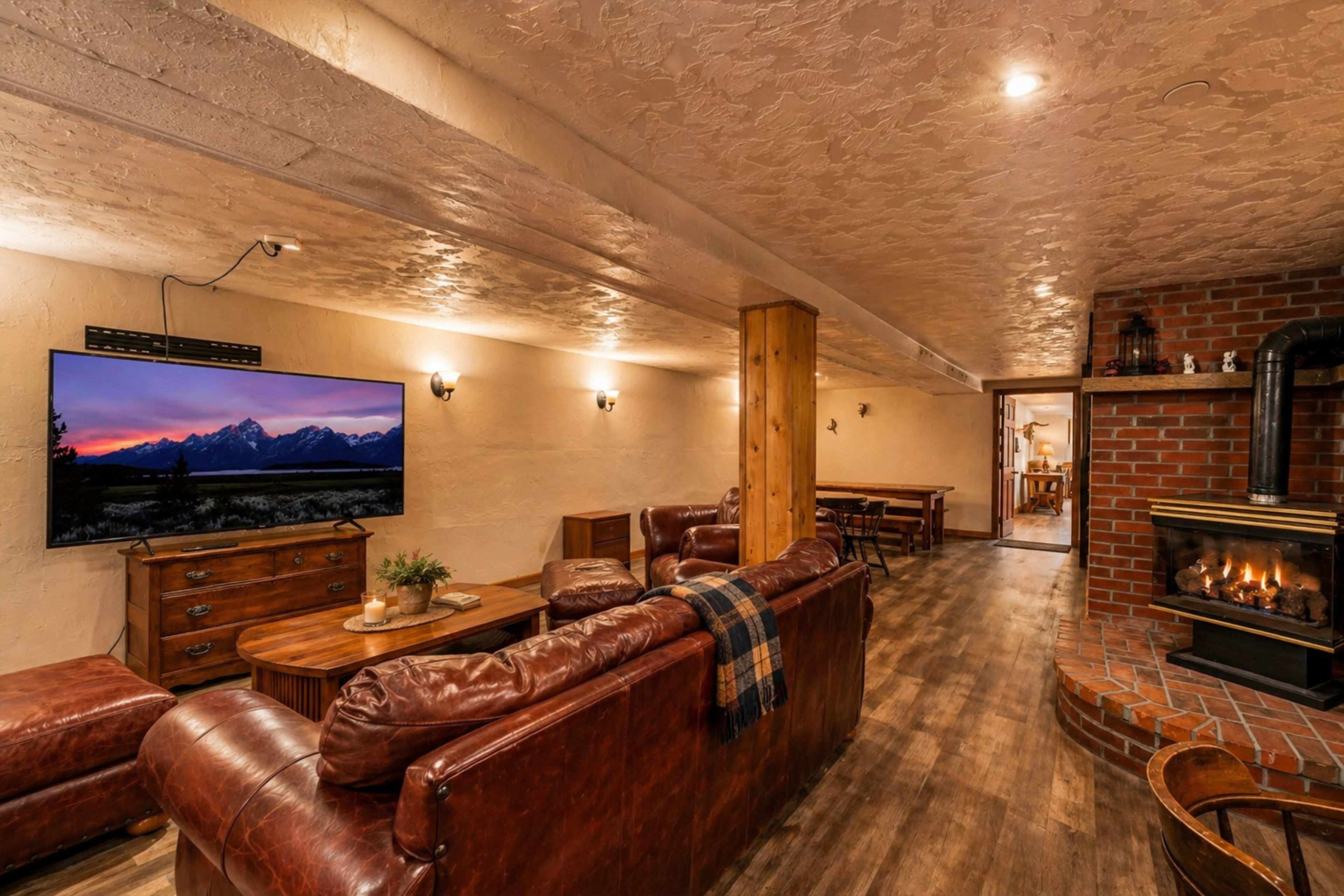 The image shows a cozy living area with leather couches, a large TV displaying a mountain landscape, and a brick fireplace.