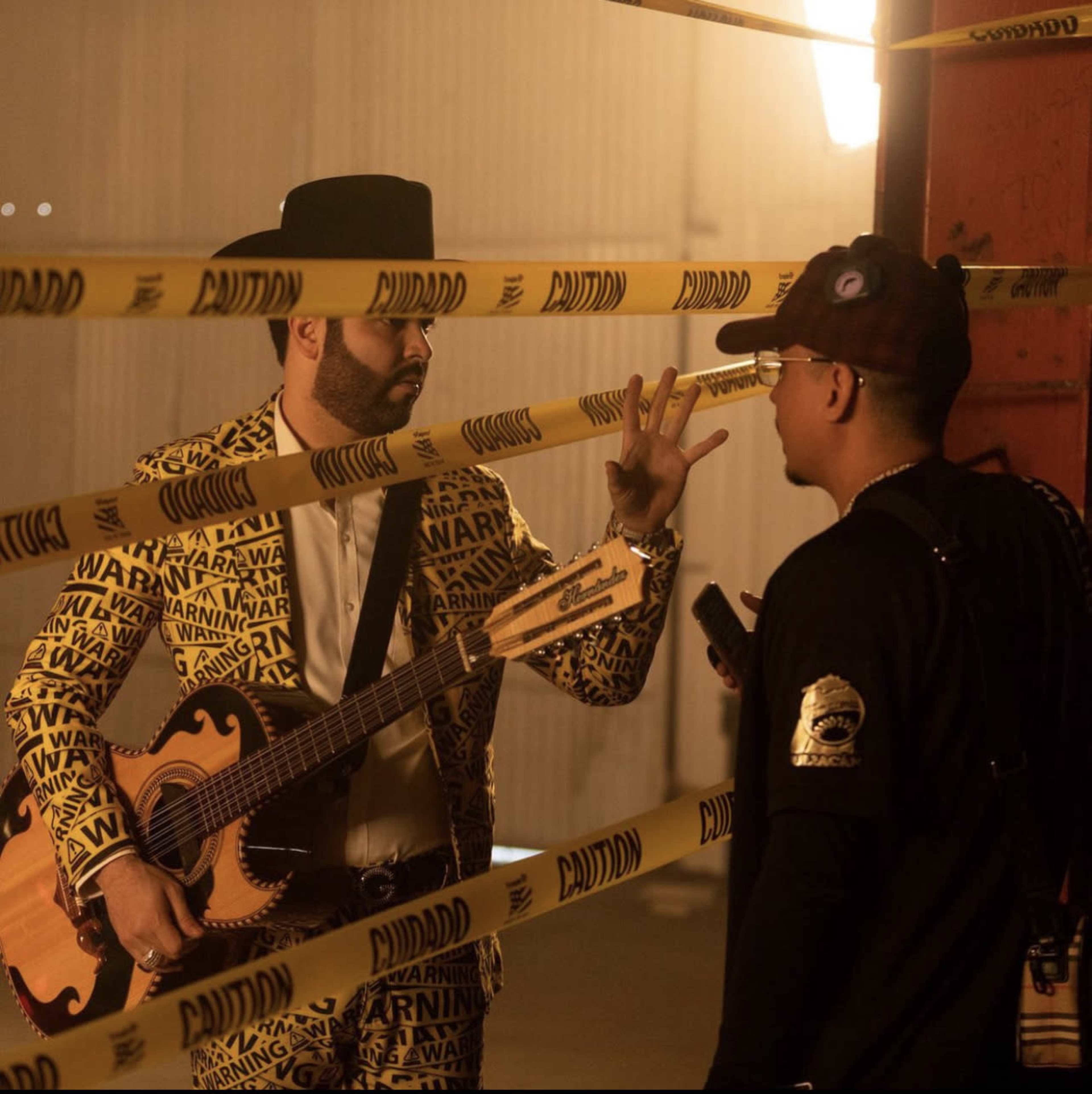 A musician in a patterned suit plays a guitar while another man interacts with him through caution tape.