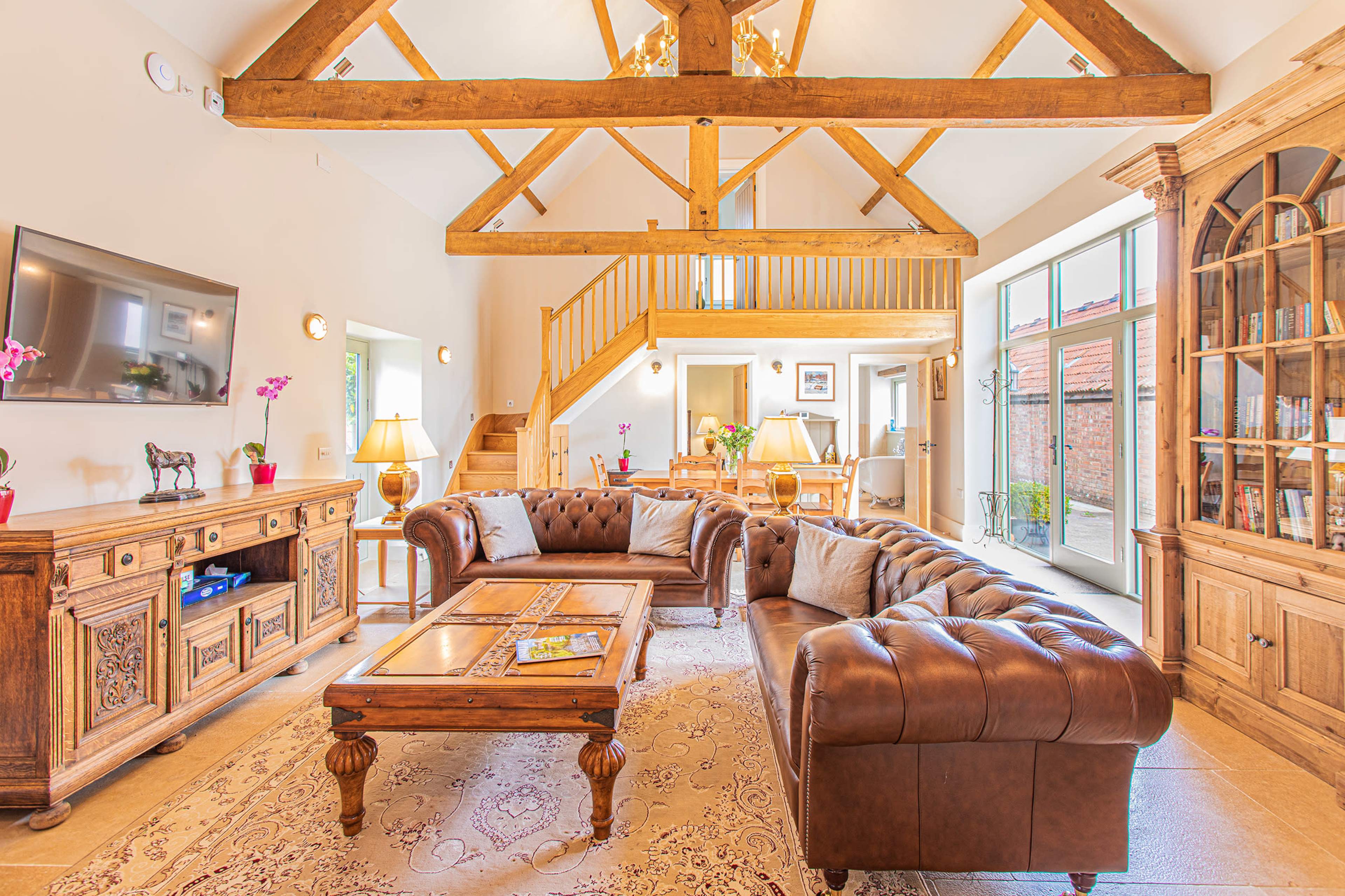 A spacious living room with a high wooden beam ceiling, a leather sectional sofa, a wooden coffee table, a large TV mounted on the wall, and natural light coming through large windows.