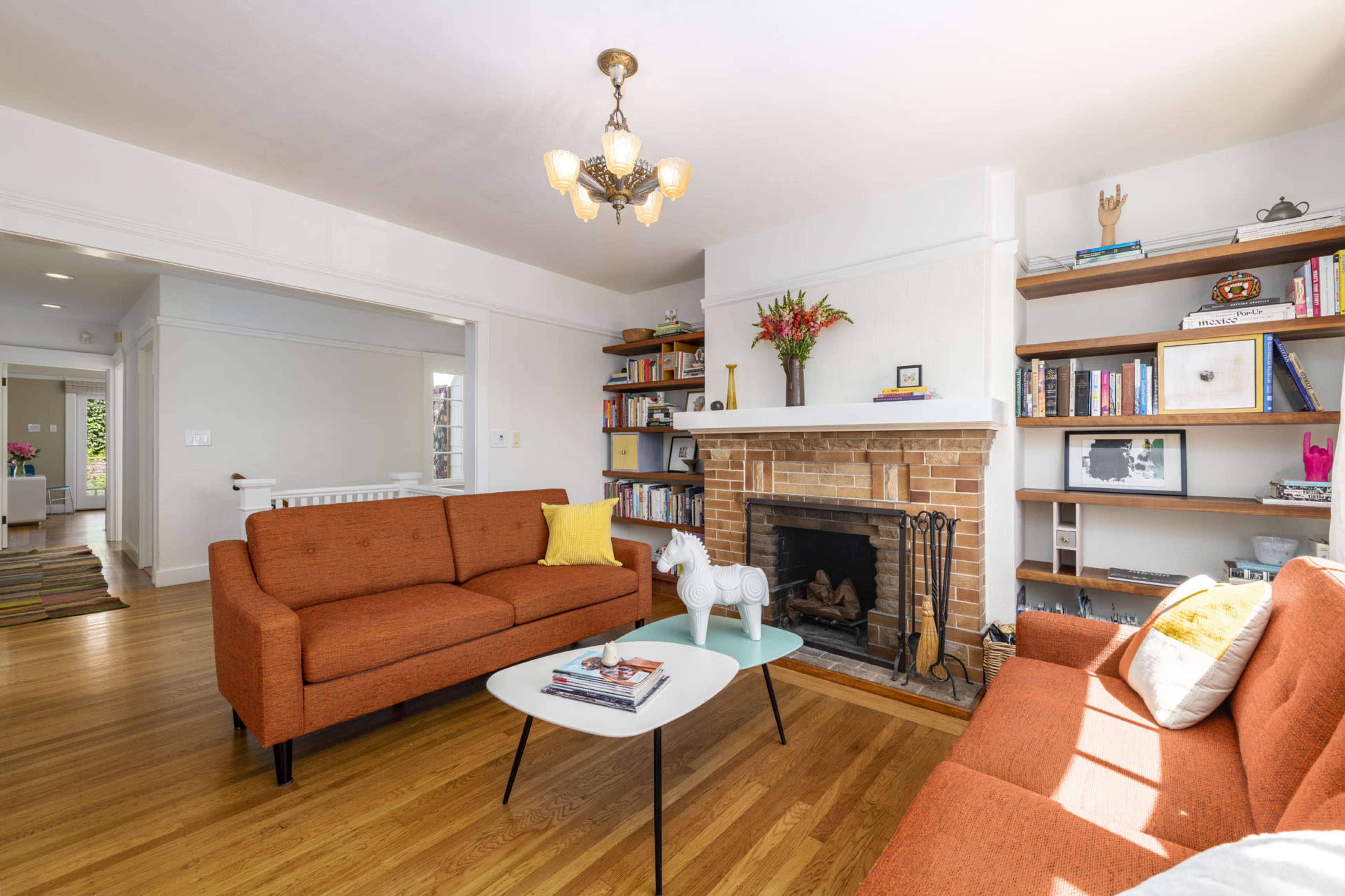 A living room with an orange upholstered sofa, a white coffee table, a brick fireplace, and shelves filled with books and decorative items.