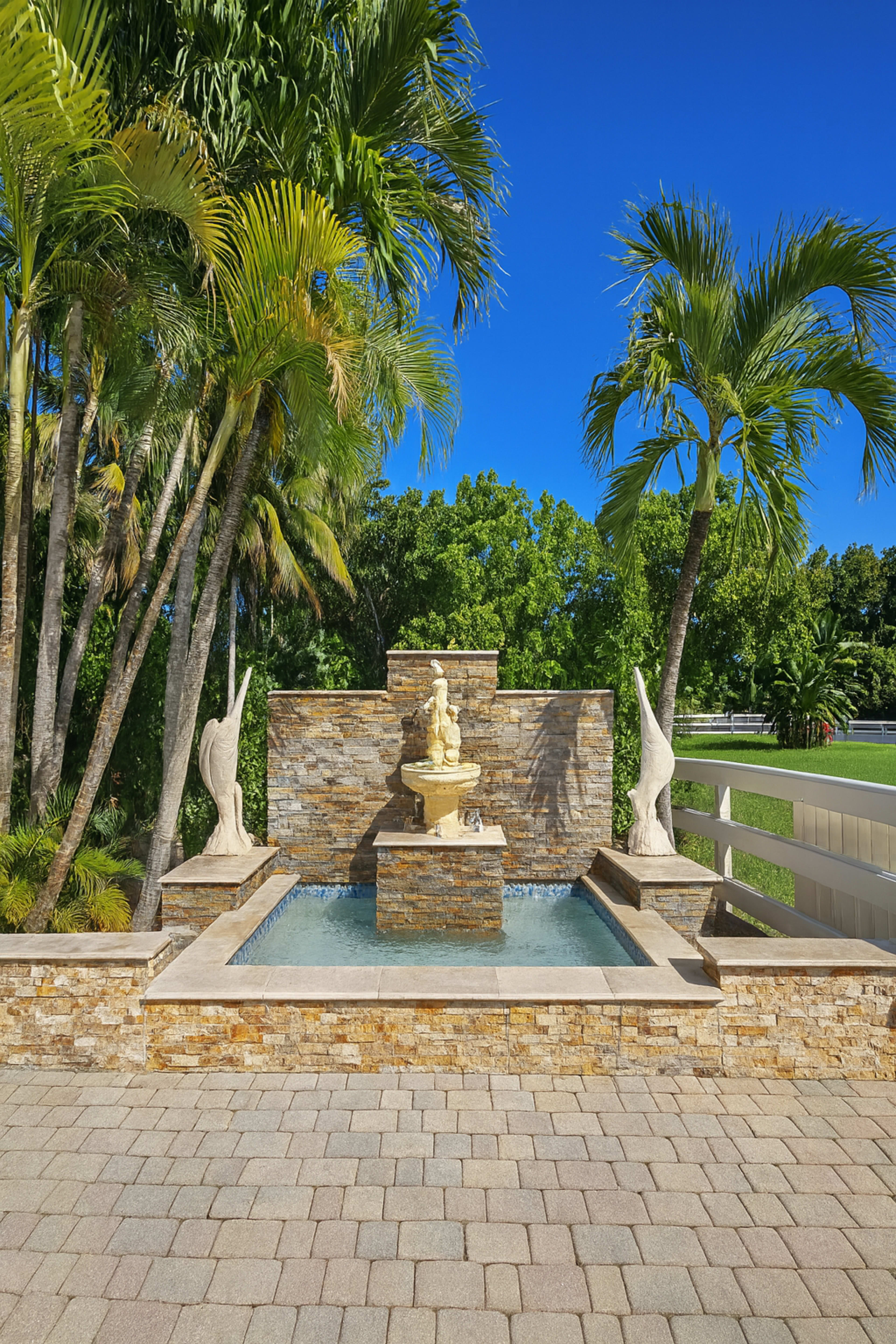 A stone fountain with a statue is set against a backdrop of palm trees and clear blue skies.