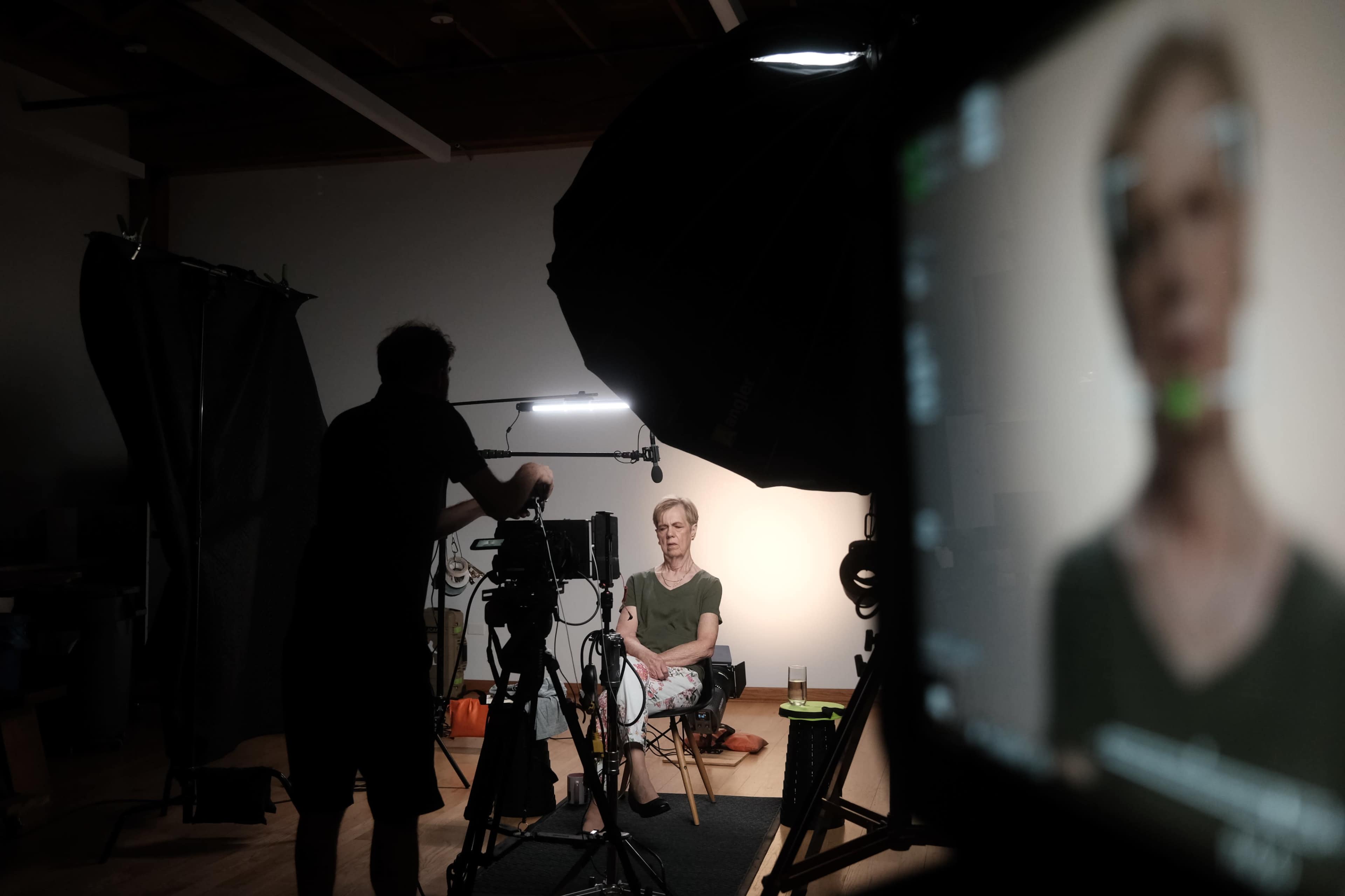 A person sits on a chair in front of a camera setup in a dimly lit studio, while another individual adjusts the camera equipment.