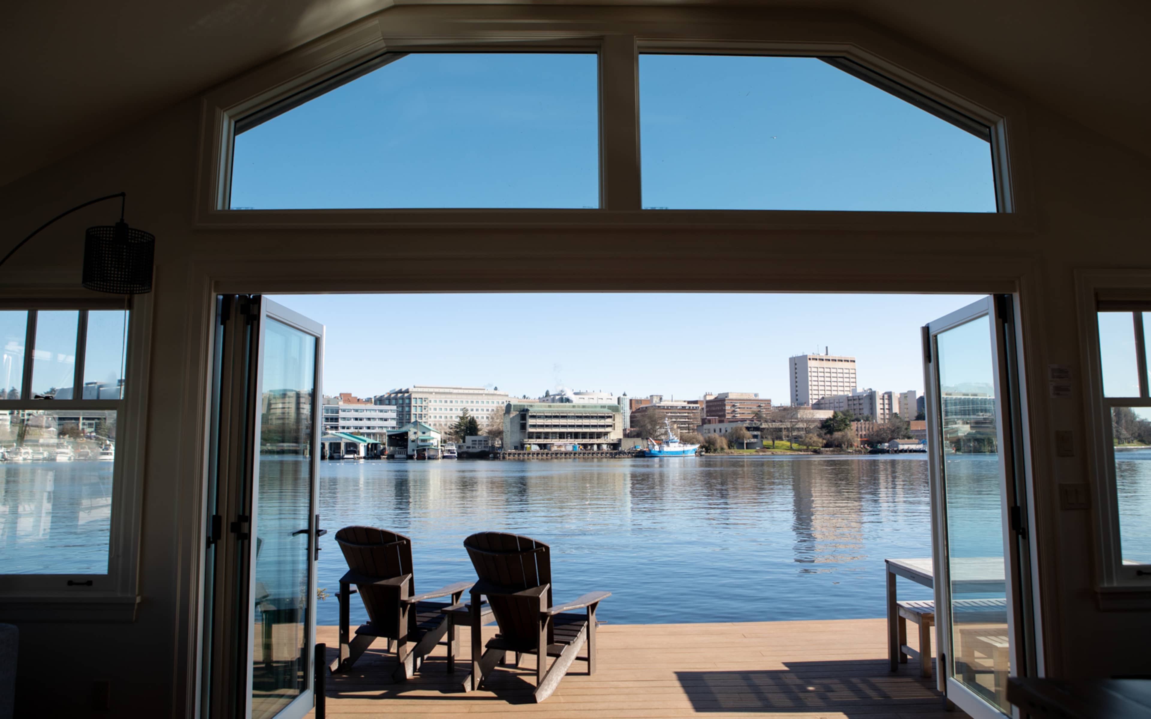 A view of a calm lake from a house, framed by open doors, with two chairs on a deck.