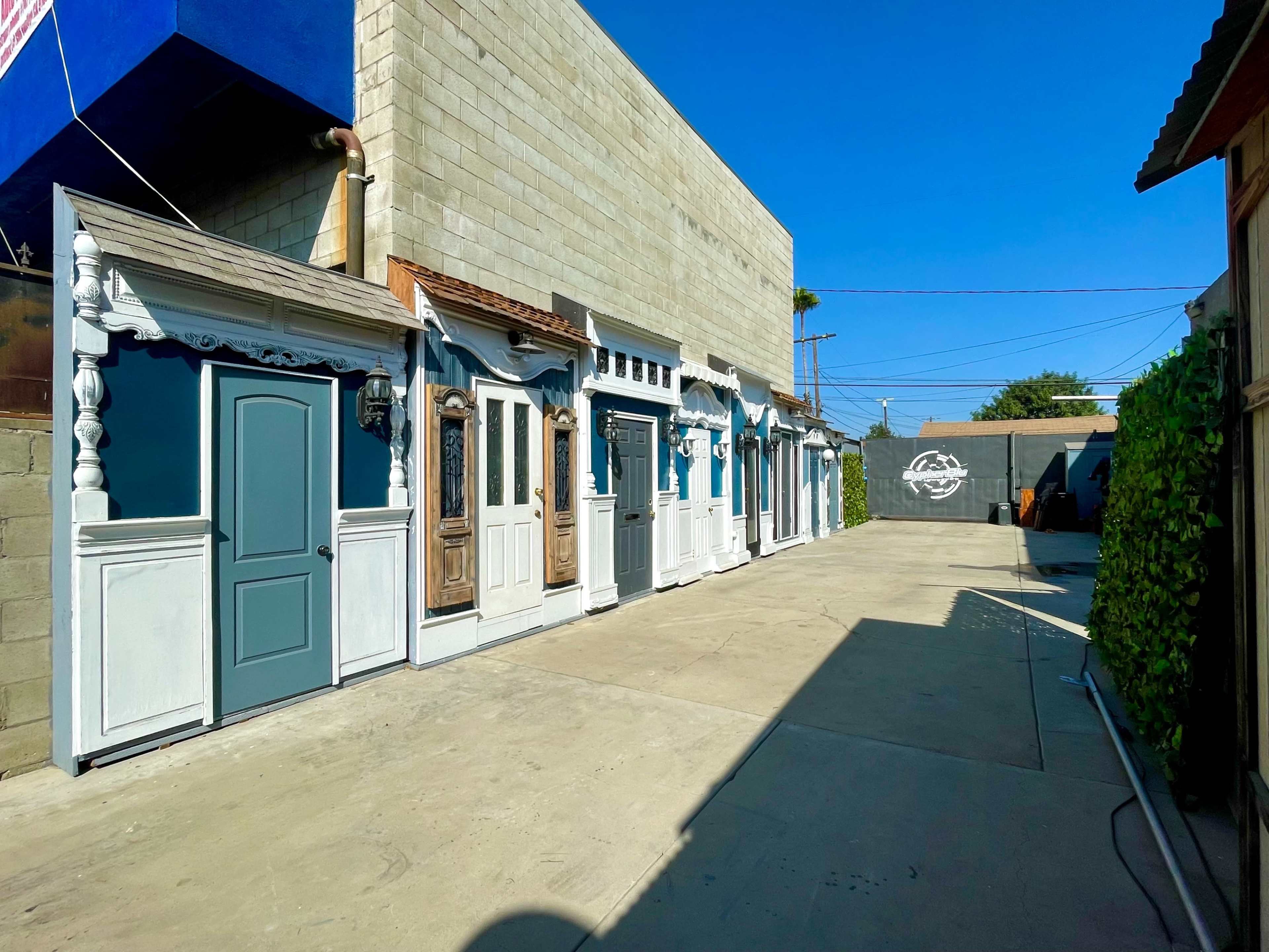 The image shows a row of decorative doors along a concrete pathway next to a gray wall, under a clear blue sky.