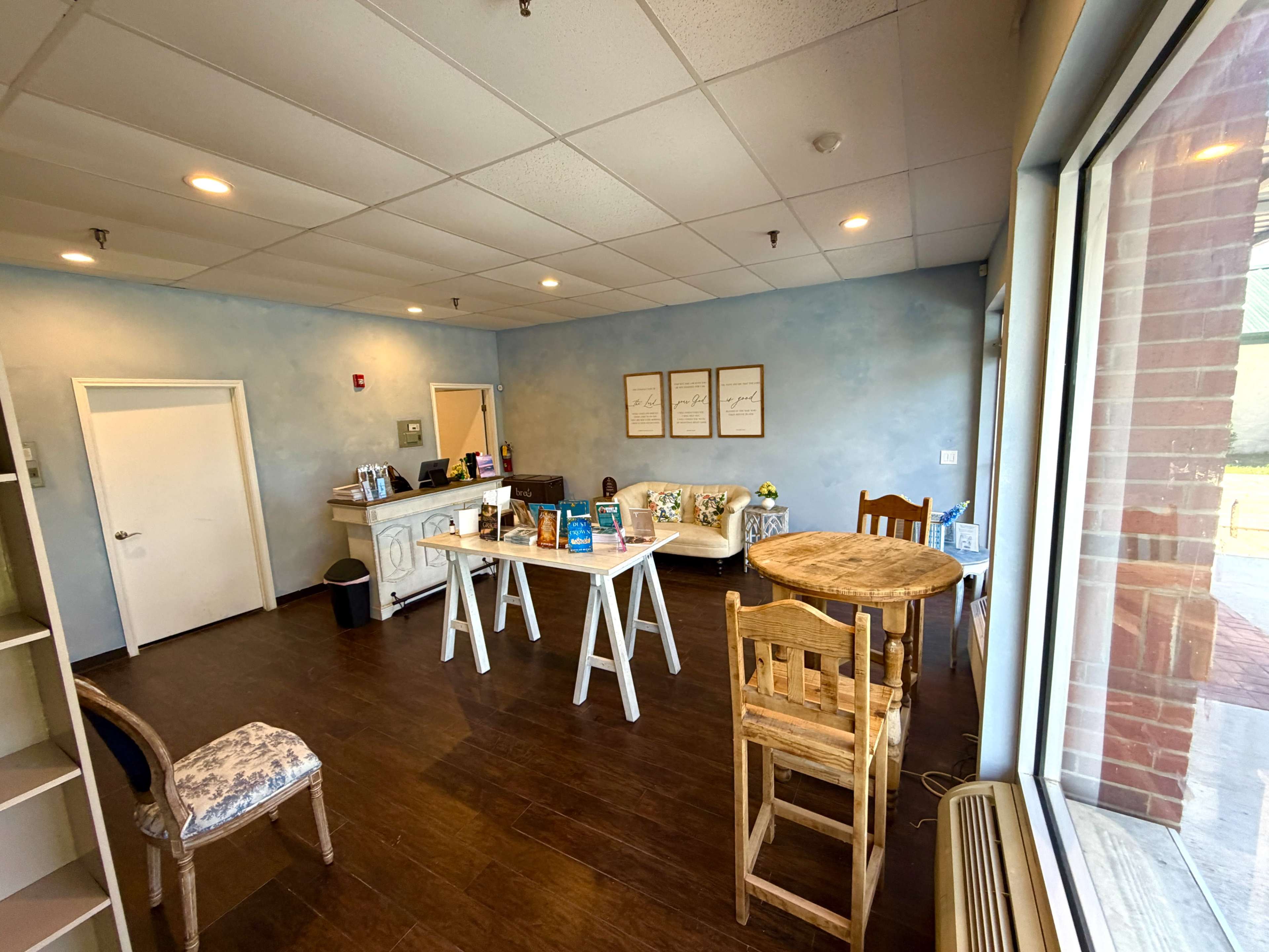 The image shows a bright, tidy room with a reception area featuring a white counter, a few tables and chairs, and decorated walls.
