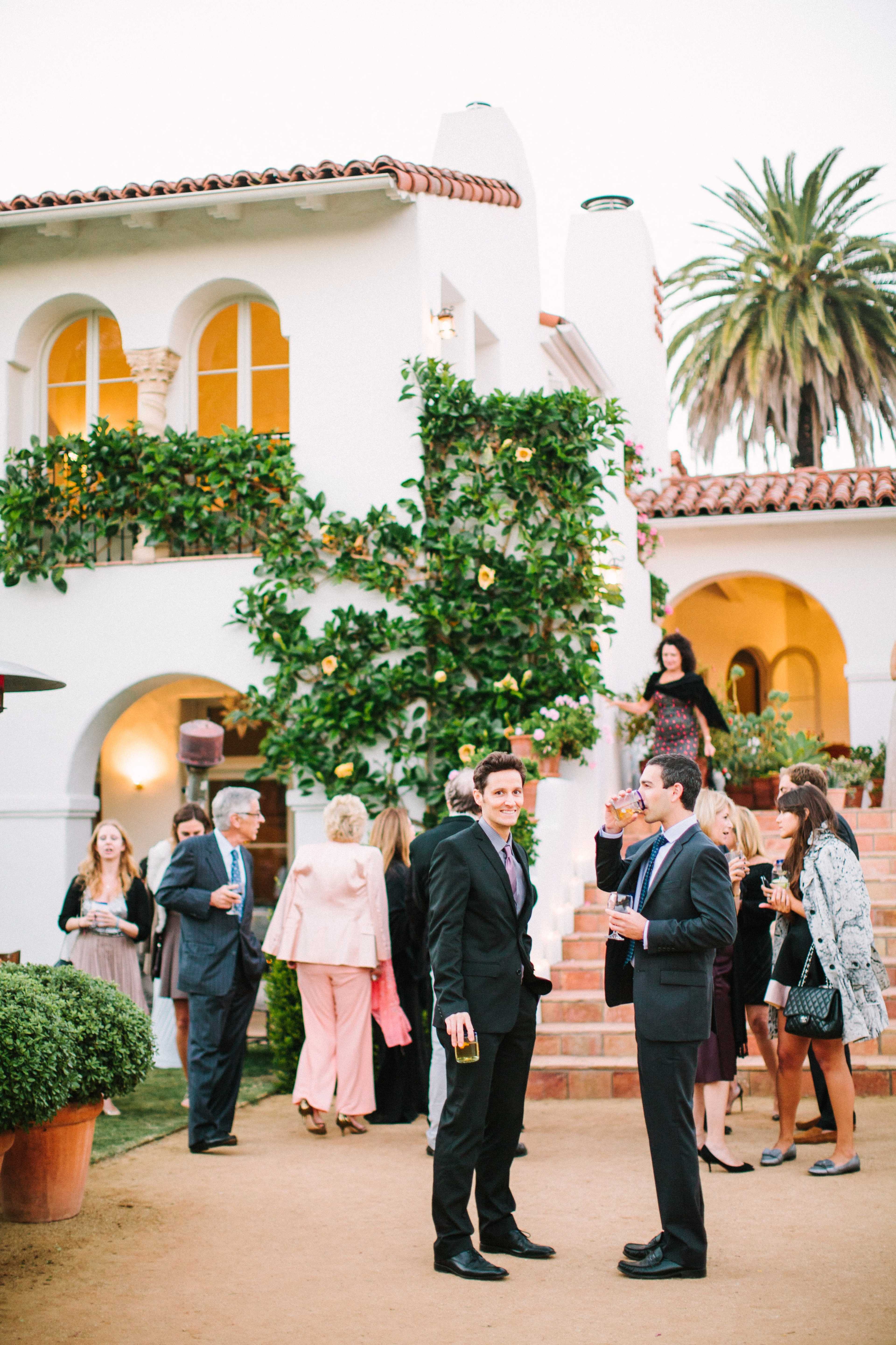 A group of people socialize on an outdoor patio in front of a stylish white building adorned with plants and palm trees.