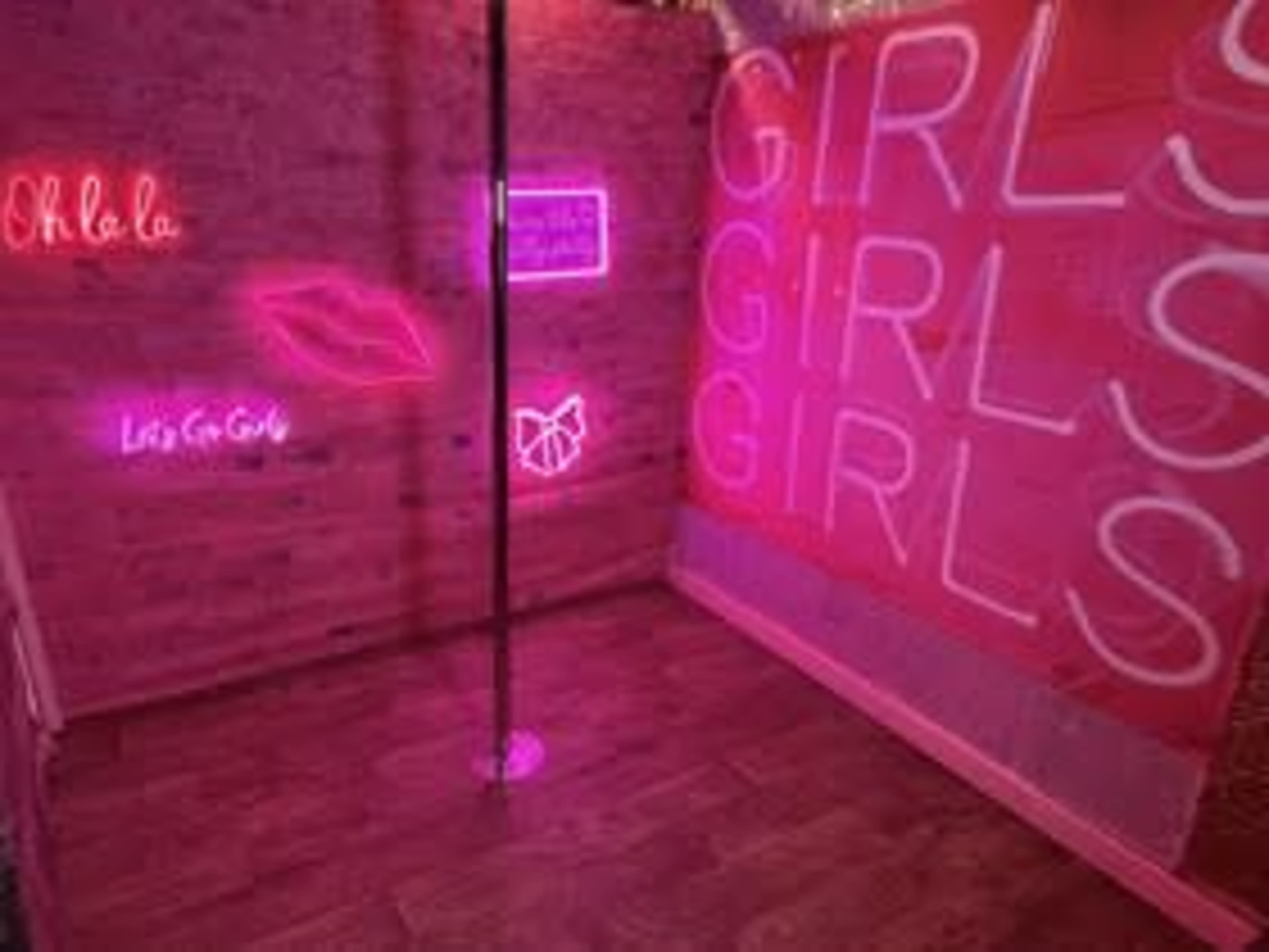 The image shows a brightly lit room featuring a dance pole, pink walls with neon signs, and a wooden floor.
