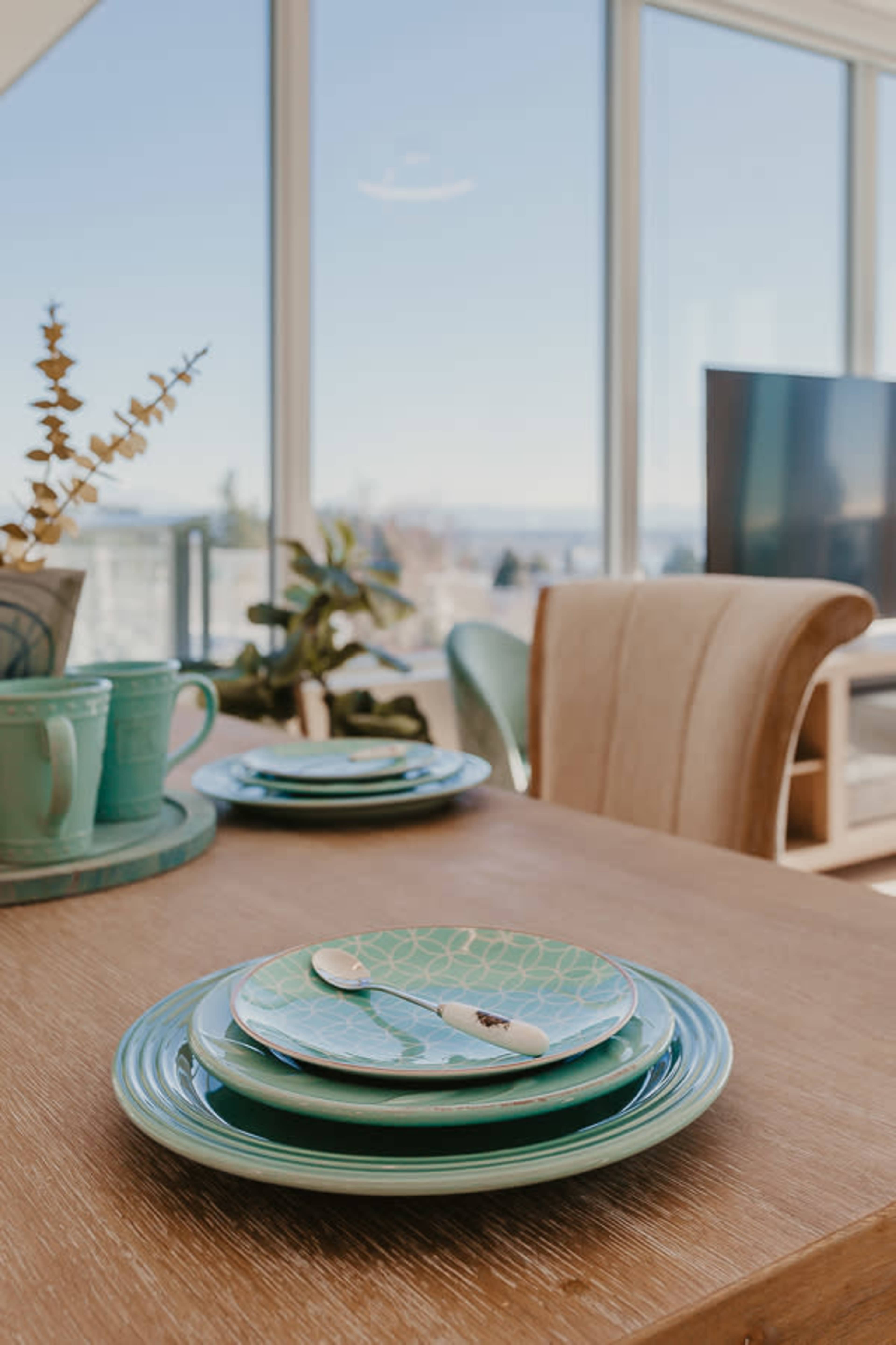 A dining table with stacked green plates and utensils is set against a backdrop of large windows offering a view of the outdoors.