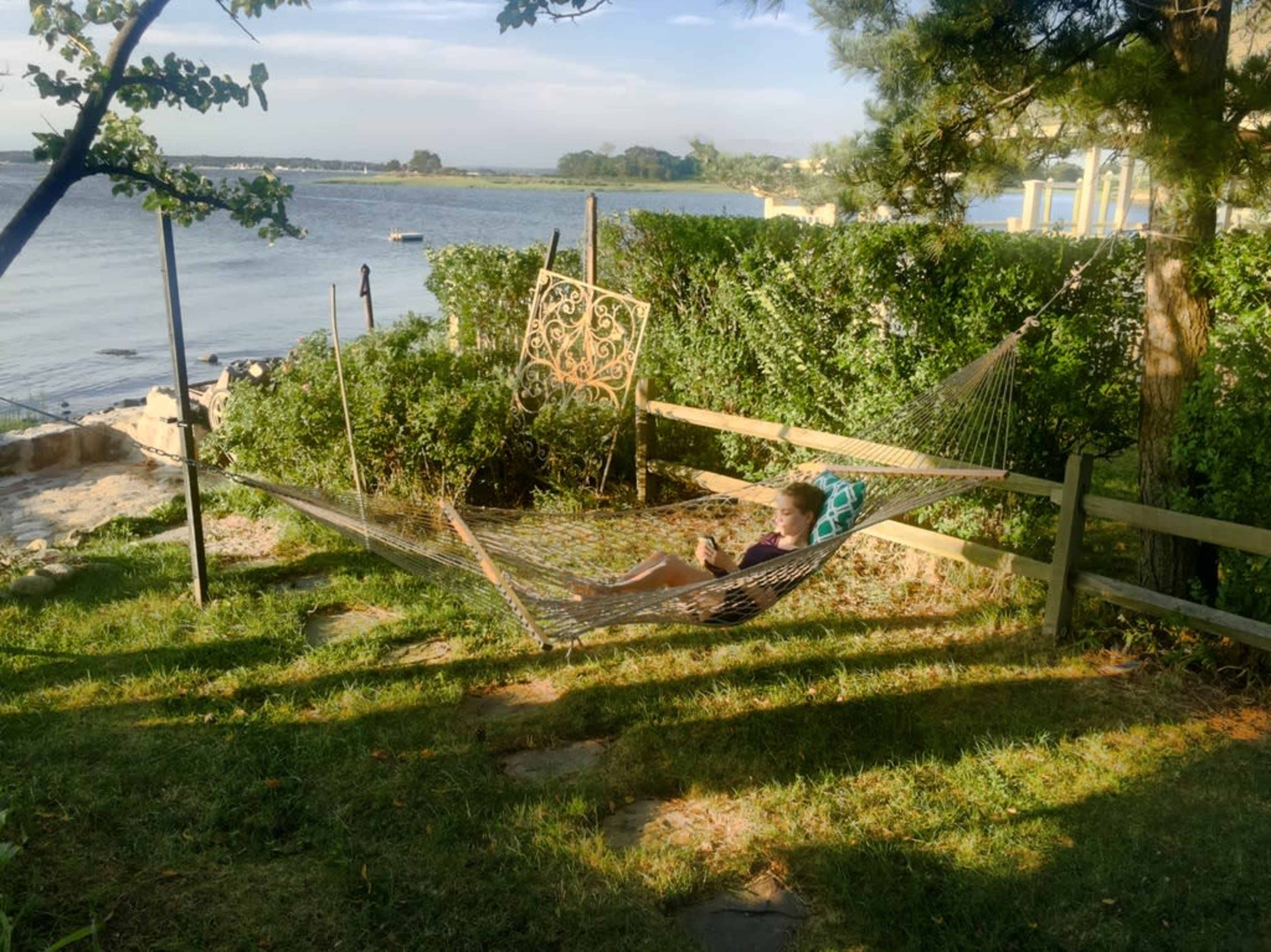 A person reclines in a hammock near a body of water, surrounded by greenery and a wooden fence.
