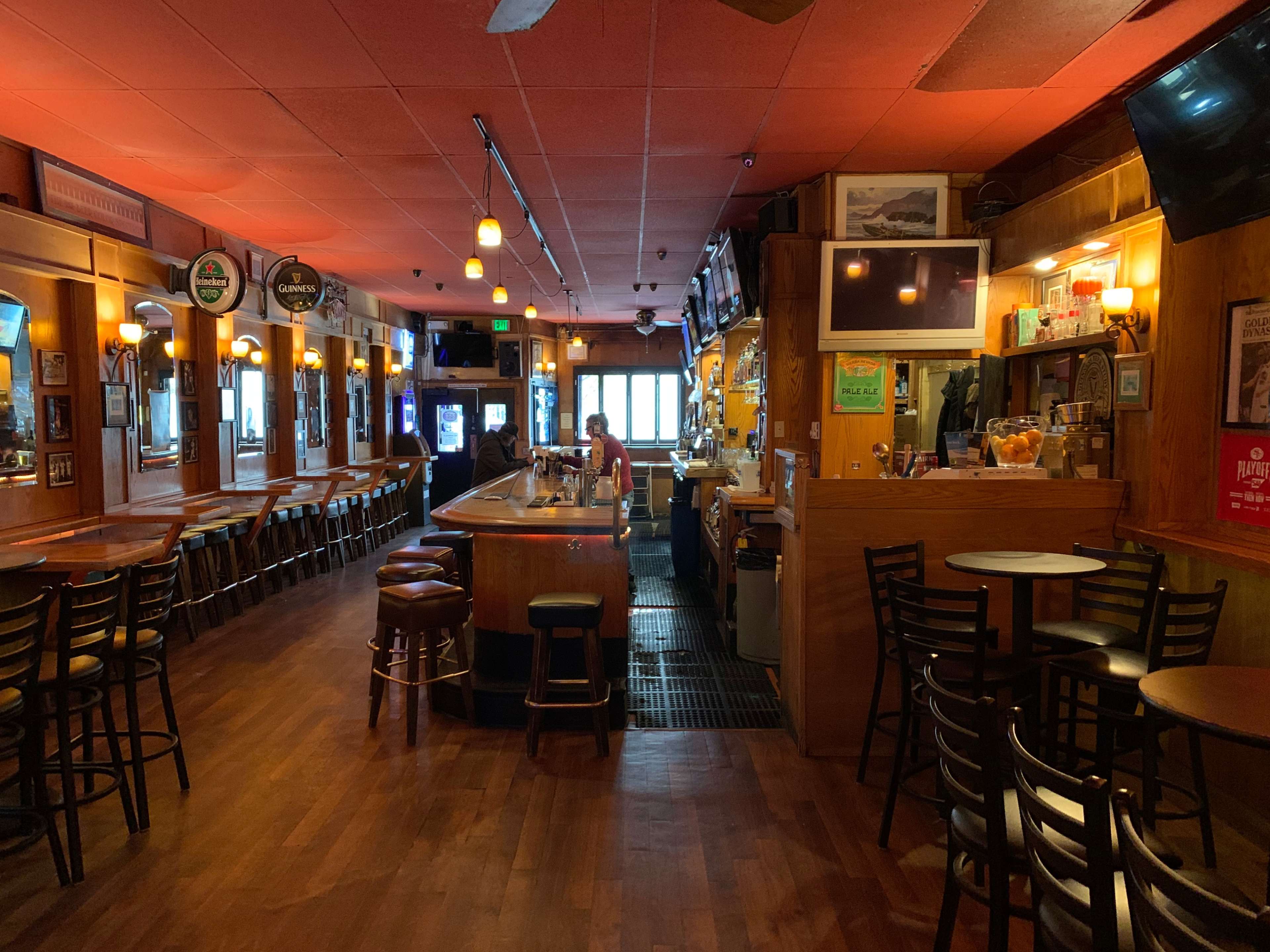 The image shows the interior of a bar with wooden furnishings, a long counter, and seating arranged around small tables.