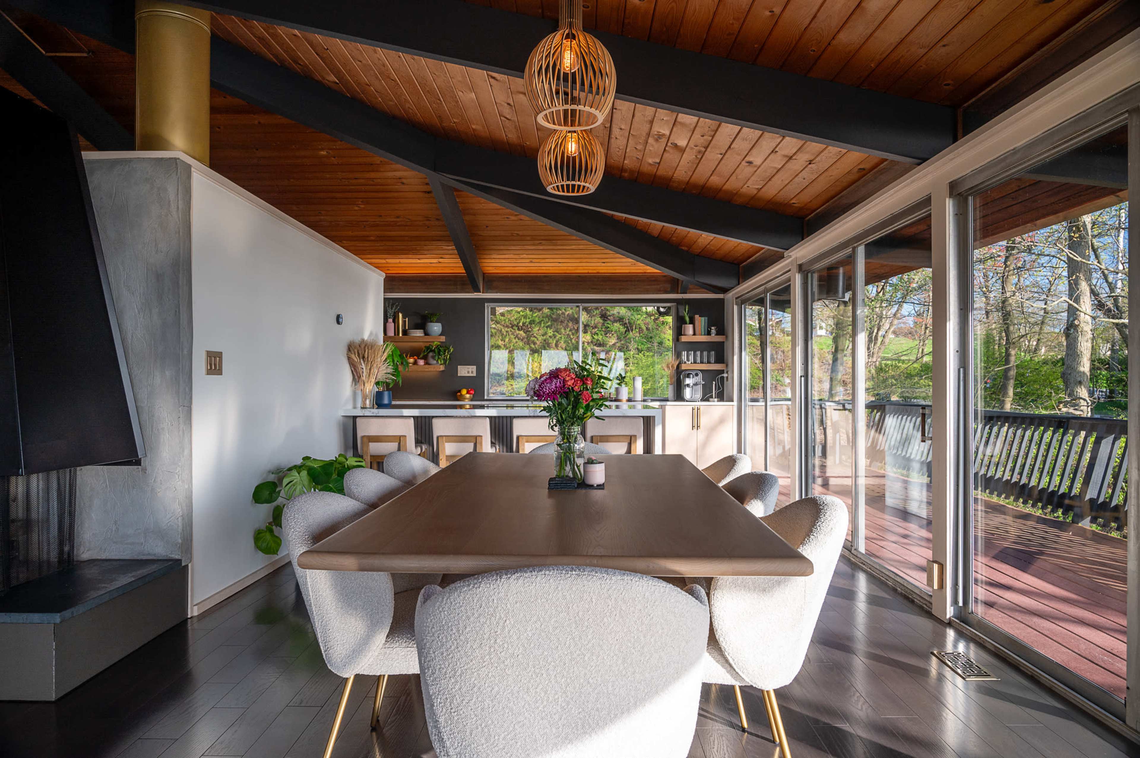 A modern dining area with a wooden table surrounded by upholstered chairs, located under a vaulted ceiling with exposed beams, and large glass doors opening to the outdoors.