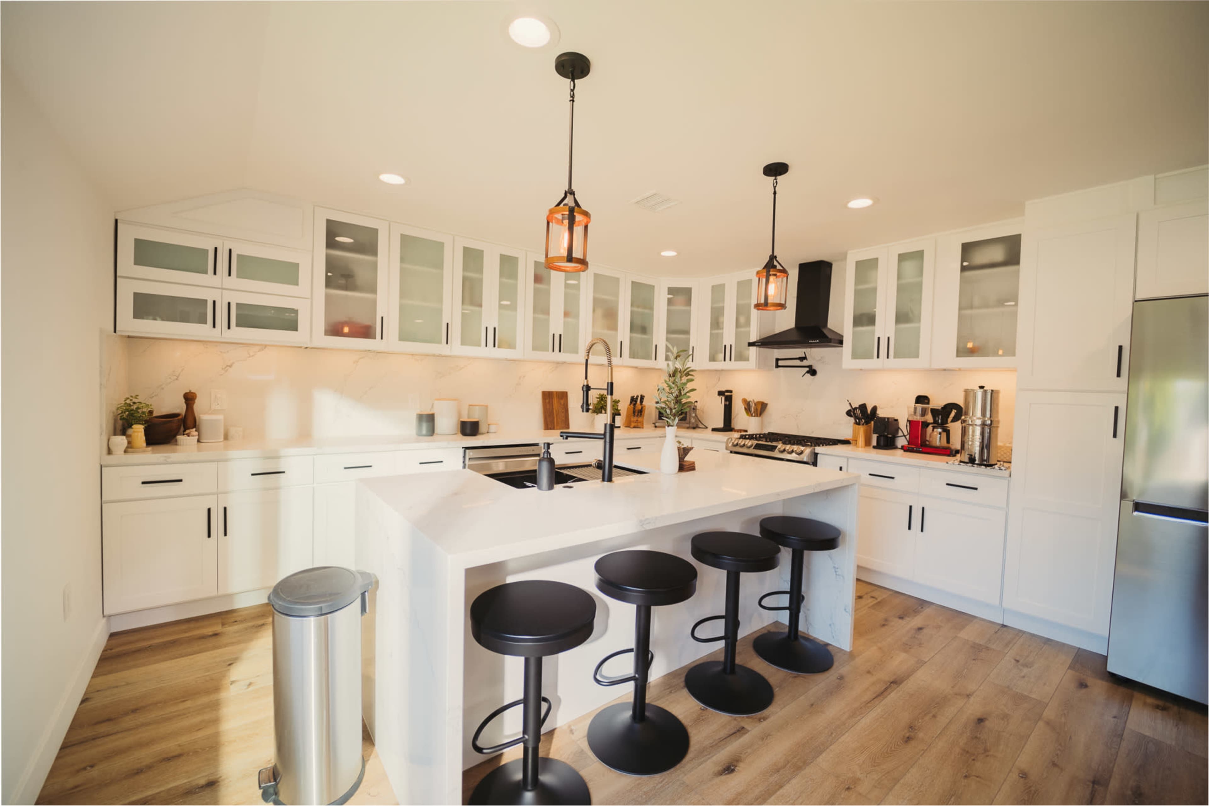 The image shows a modern kitchen featuring white cabinets, a central island with four black stools, and stainless steel appliances.