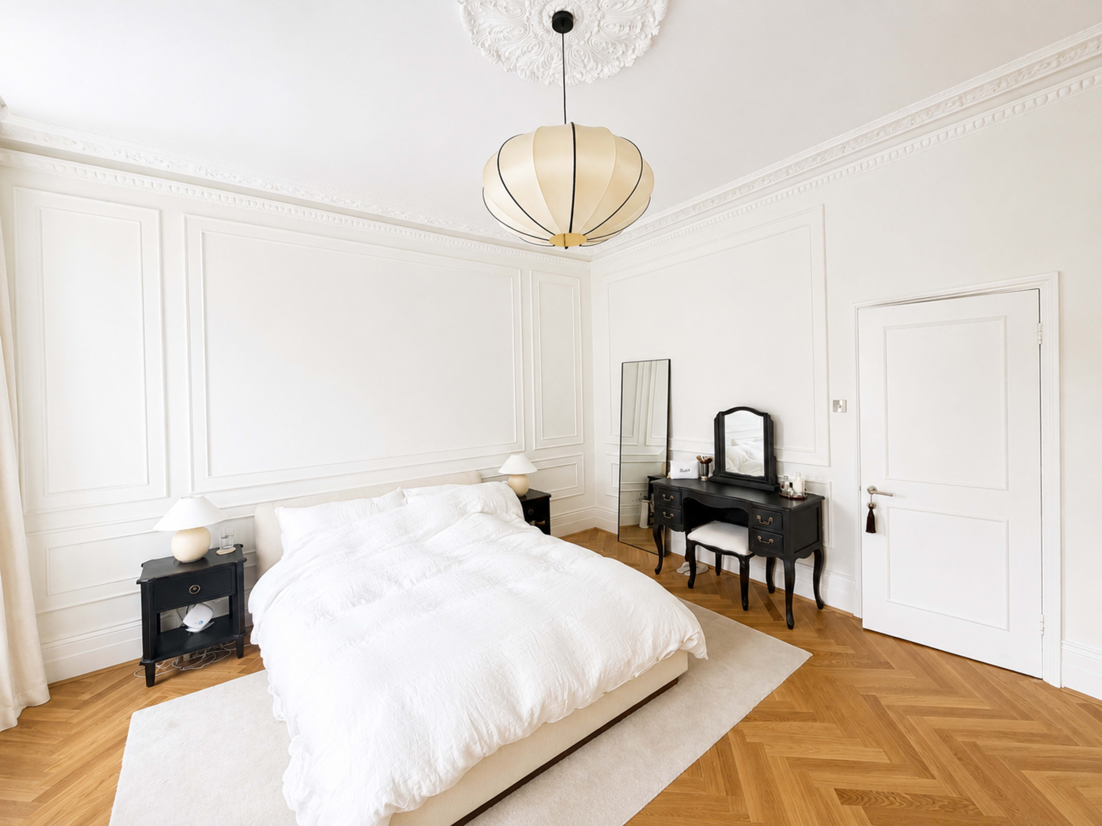 The image shows a bright, minimalist bedroom with a large bed, a vanity table with a mirror, and decorative wall molding.