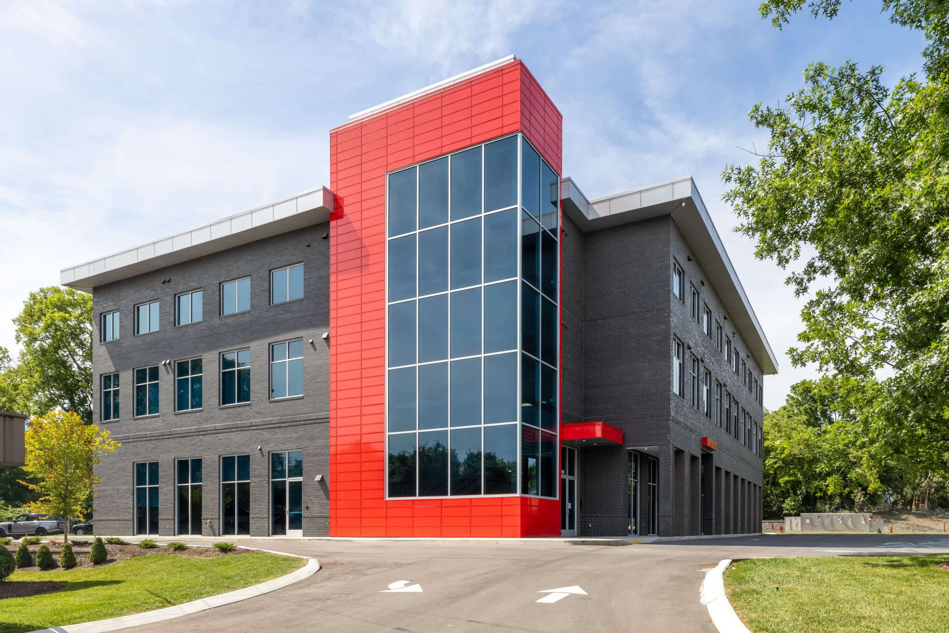 The image shows a modern three-story building with a red-accented facade and large glass windows, set on a paved driveway.