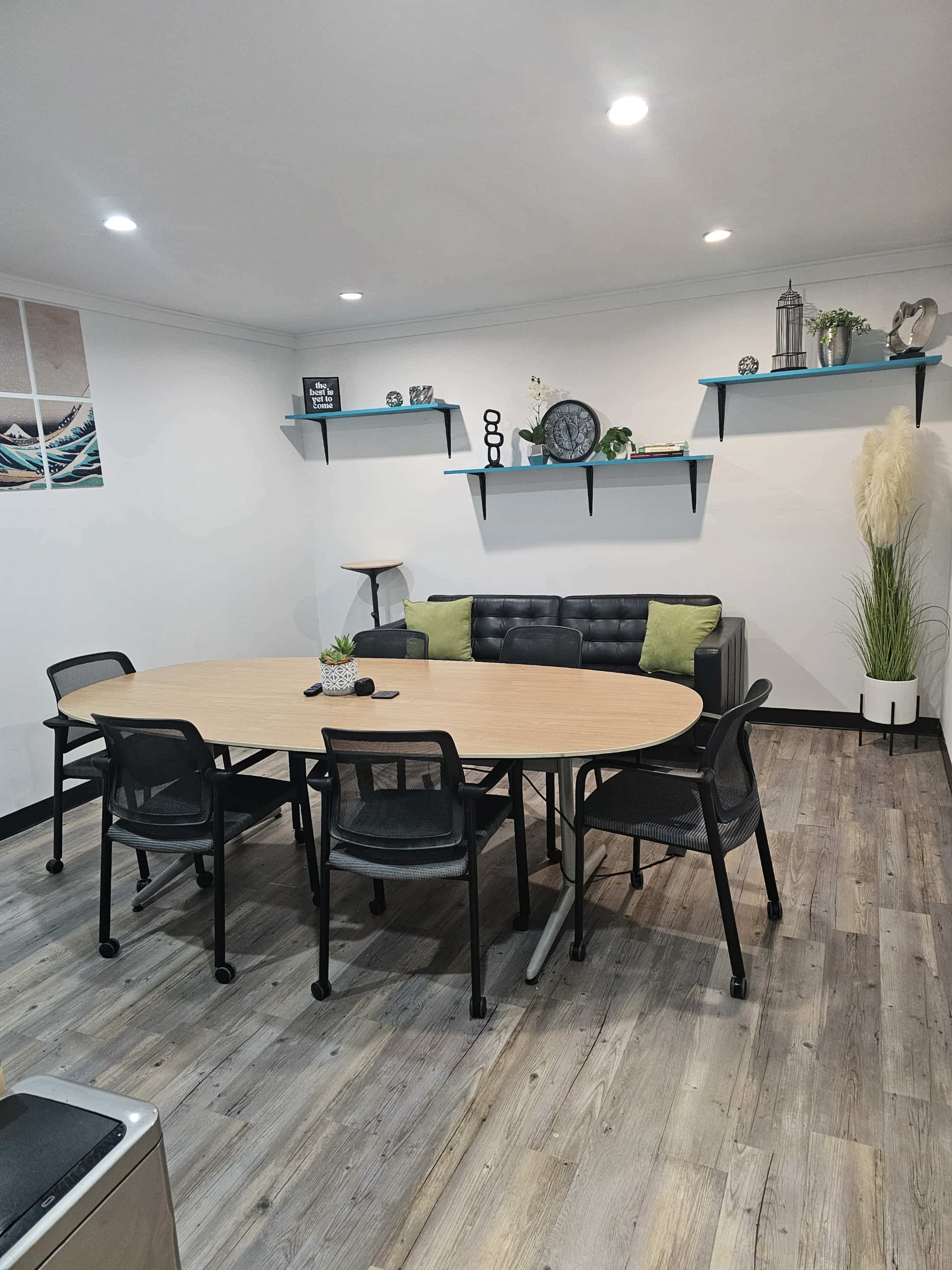 The image shows a small meeting room with a round table surrounded by black chairs, a black couch with green cushions, and shelves displaying decorative items on the walls.
