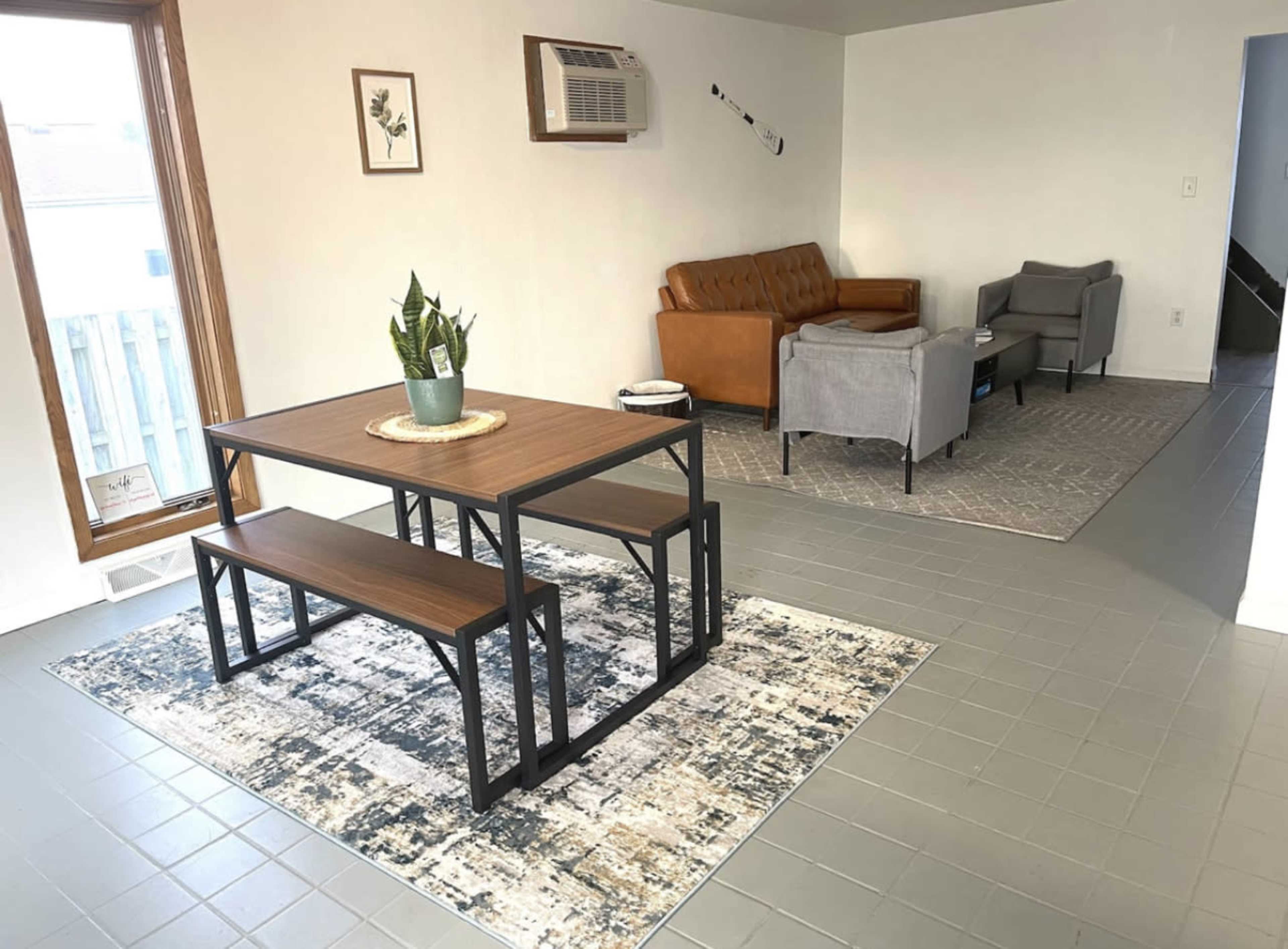 A modern living space with a dining table and benches in the foreground and a seating area featuring a brown couch and gray chairs in the background.