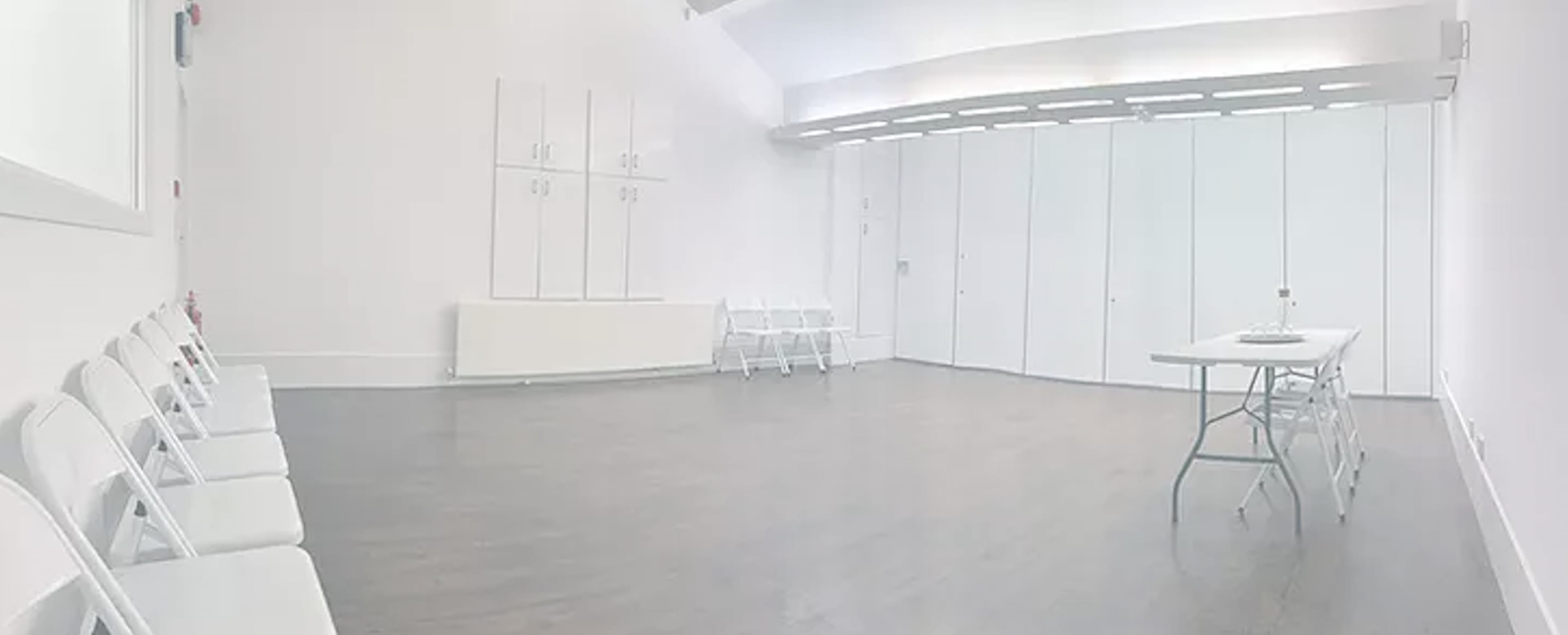 A spacious, minimally furnished room features white walls, a wooden floor, a table set for a small gathering, and several folding chairs arranged along one side.