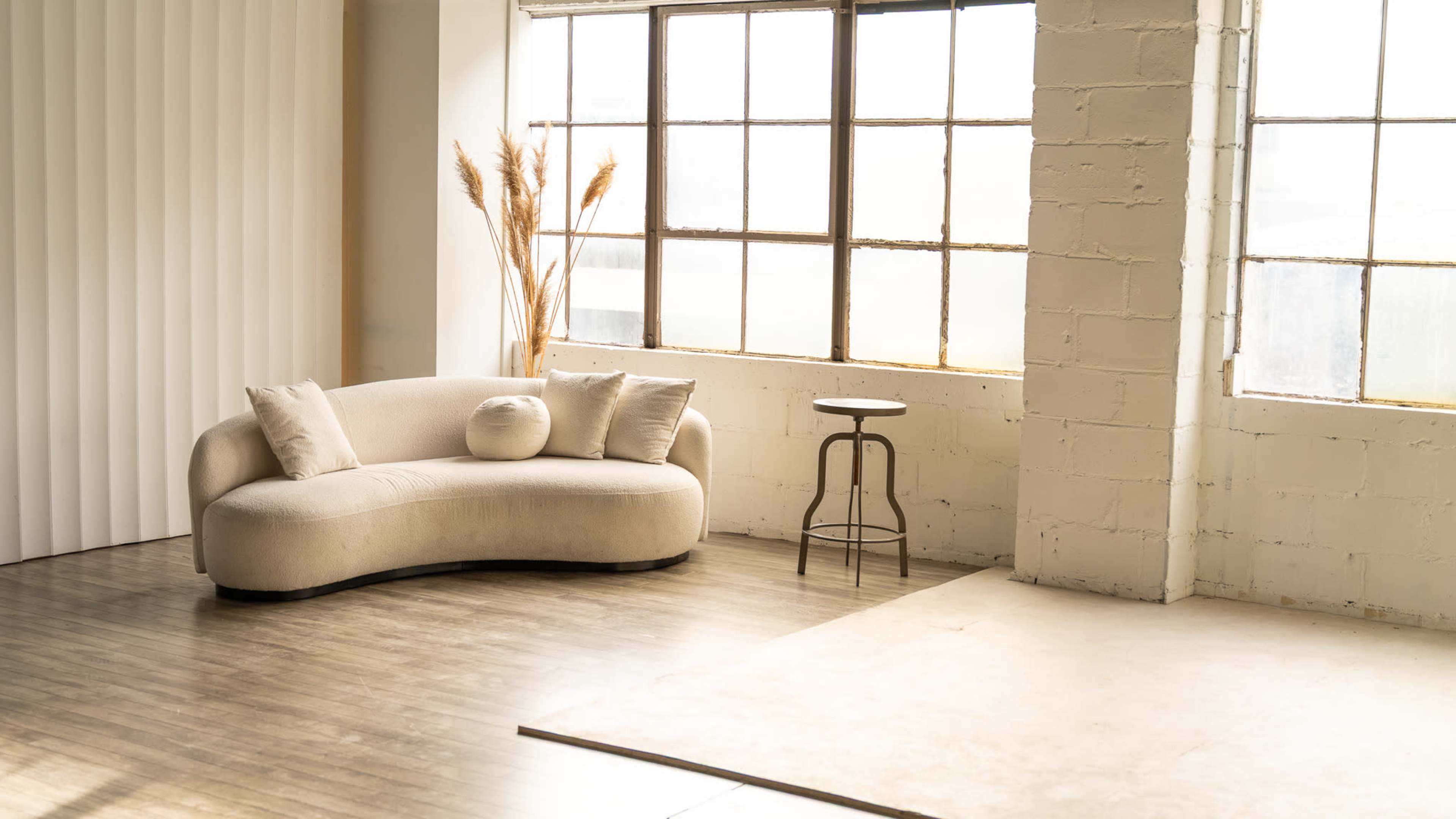 The image displays a modern, curved beige sofa next to a circular wooden table in a bright, minimalist room with large windows.