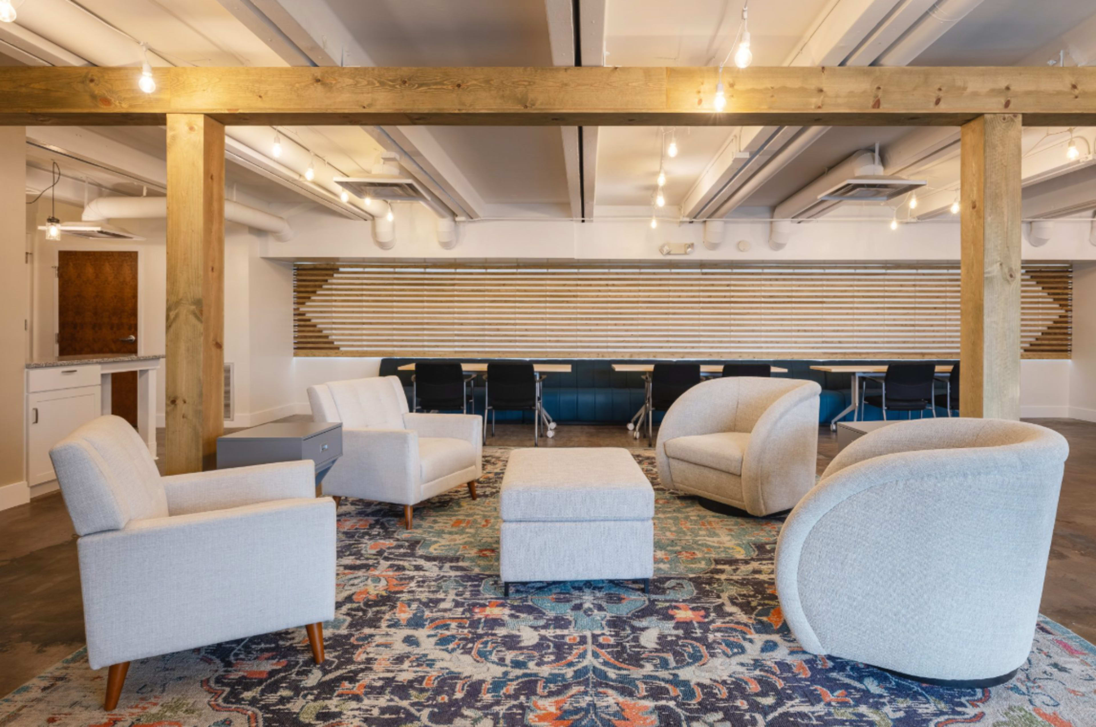 The image shows a modern lounge area with four upholstered chairs arranged around a central ottoman, set on a decorative rug under pendant lights in a spacious room.