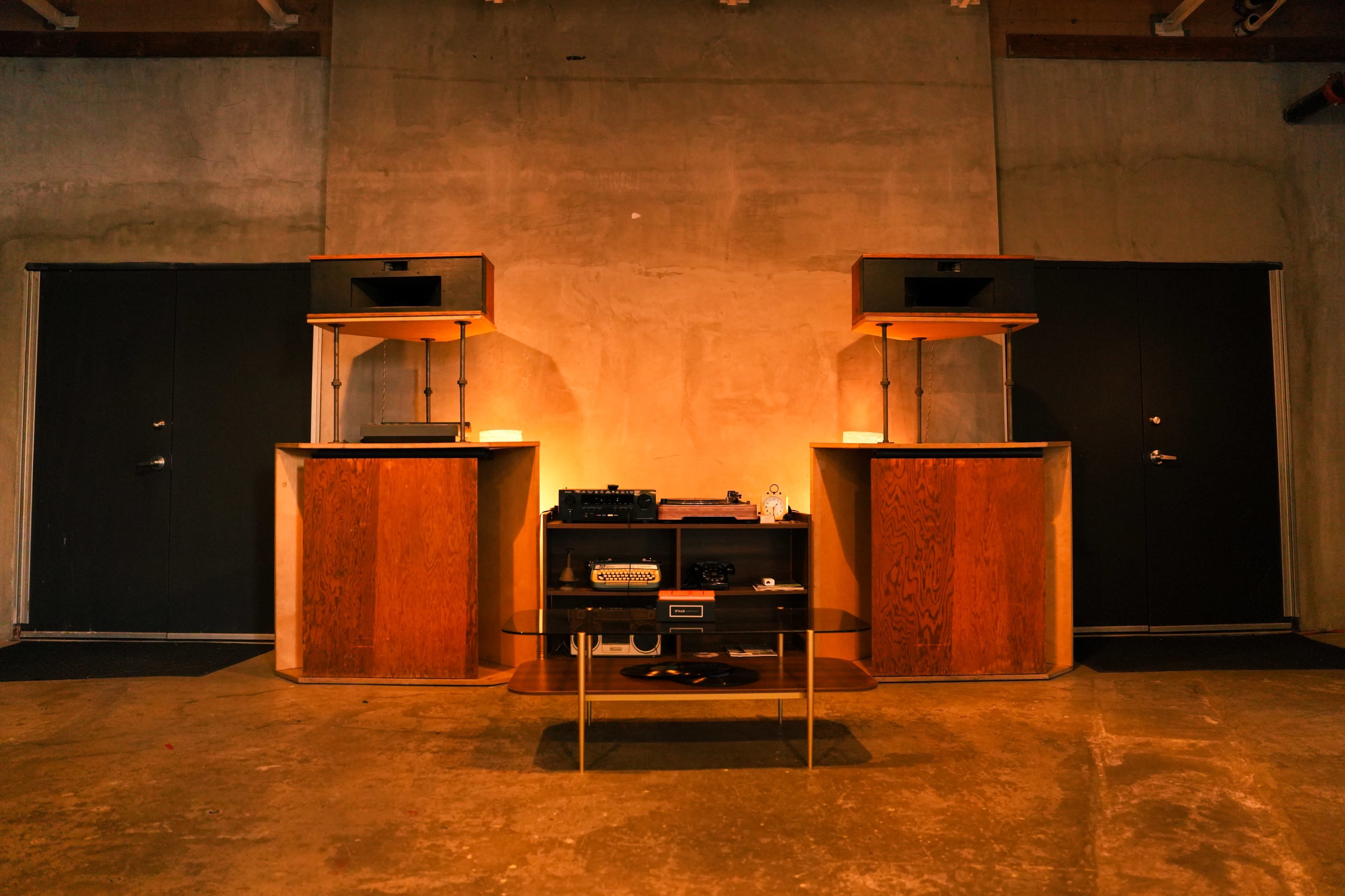 The image shows a minimalist interior with two wooden speaker stands flanking a low-profile audio setup on a table, against a concrete wall and two dark doors.