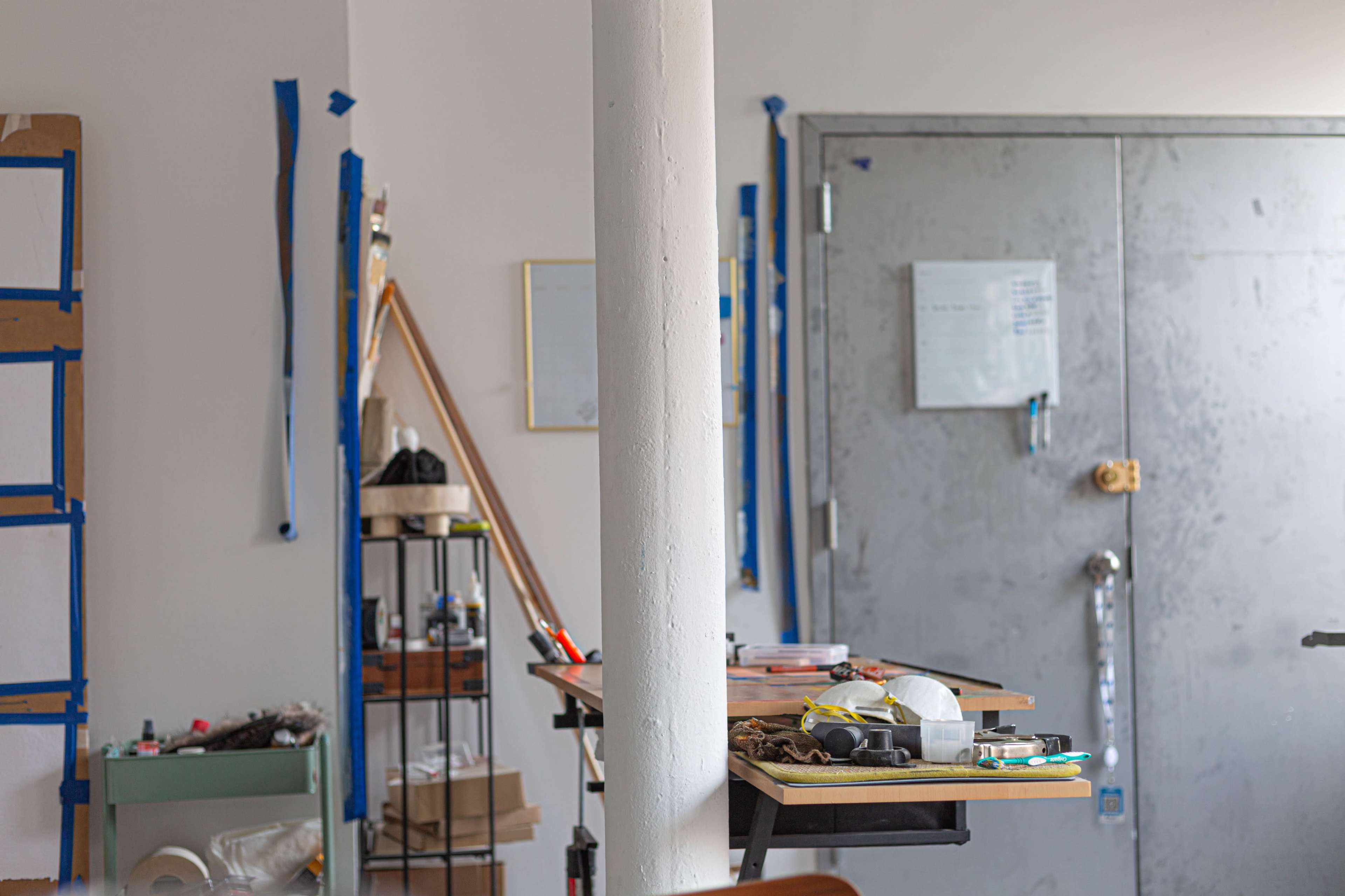 The image shows a cluttered workspace with various tools and materials on a table, a white column in the foreground, and a metallic door in the background.