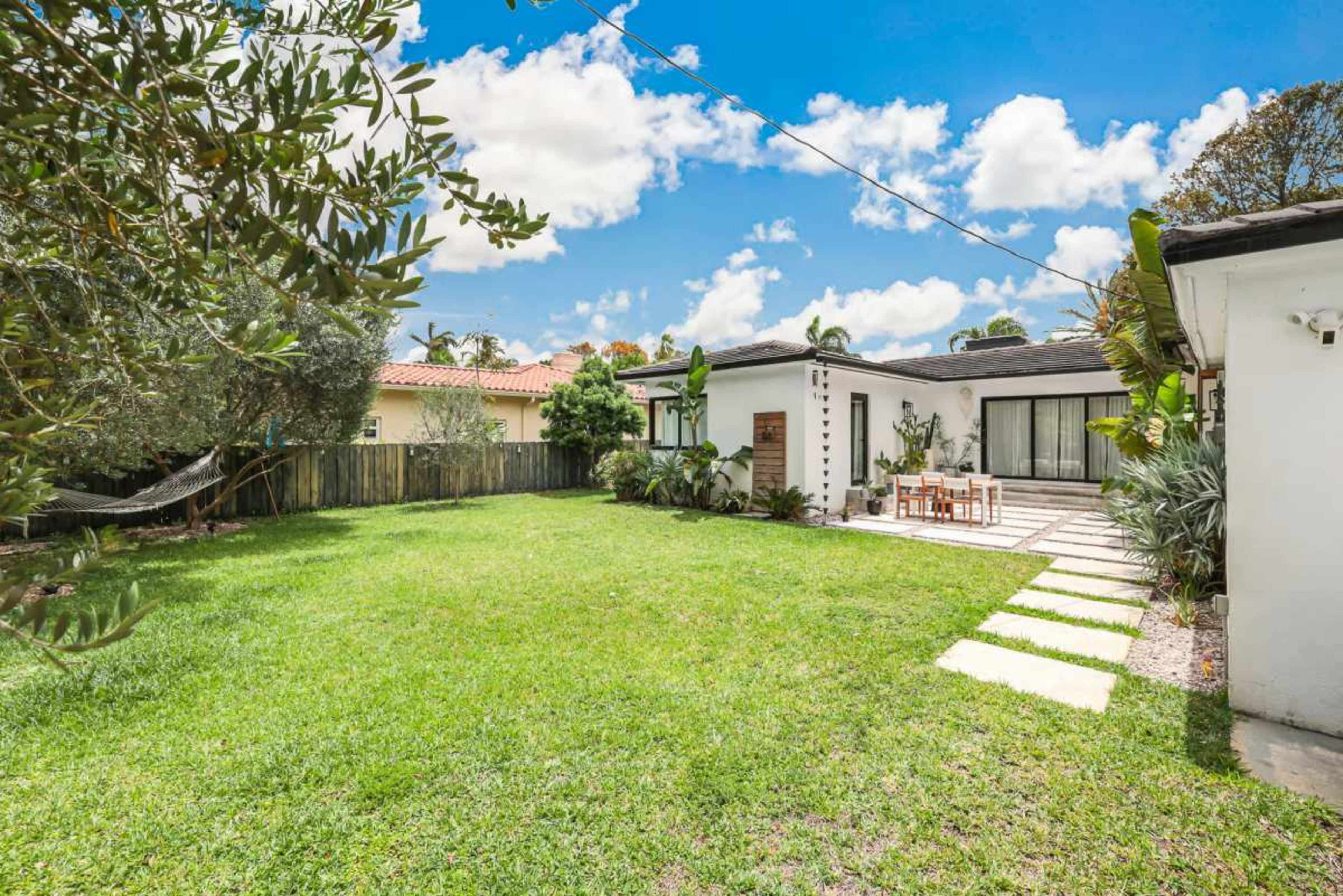 The image shows a spacious backyard with a well-maintained lawn, a patio area with a dining table, and surrounding greenery.