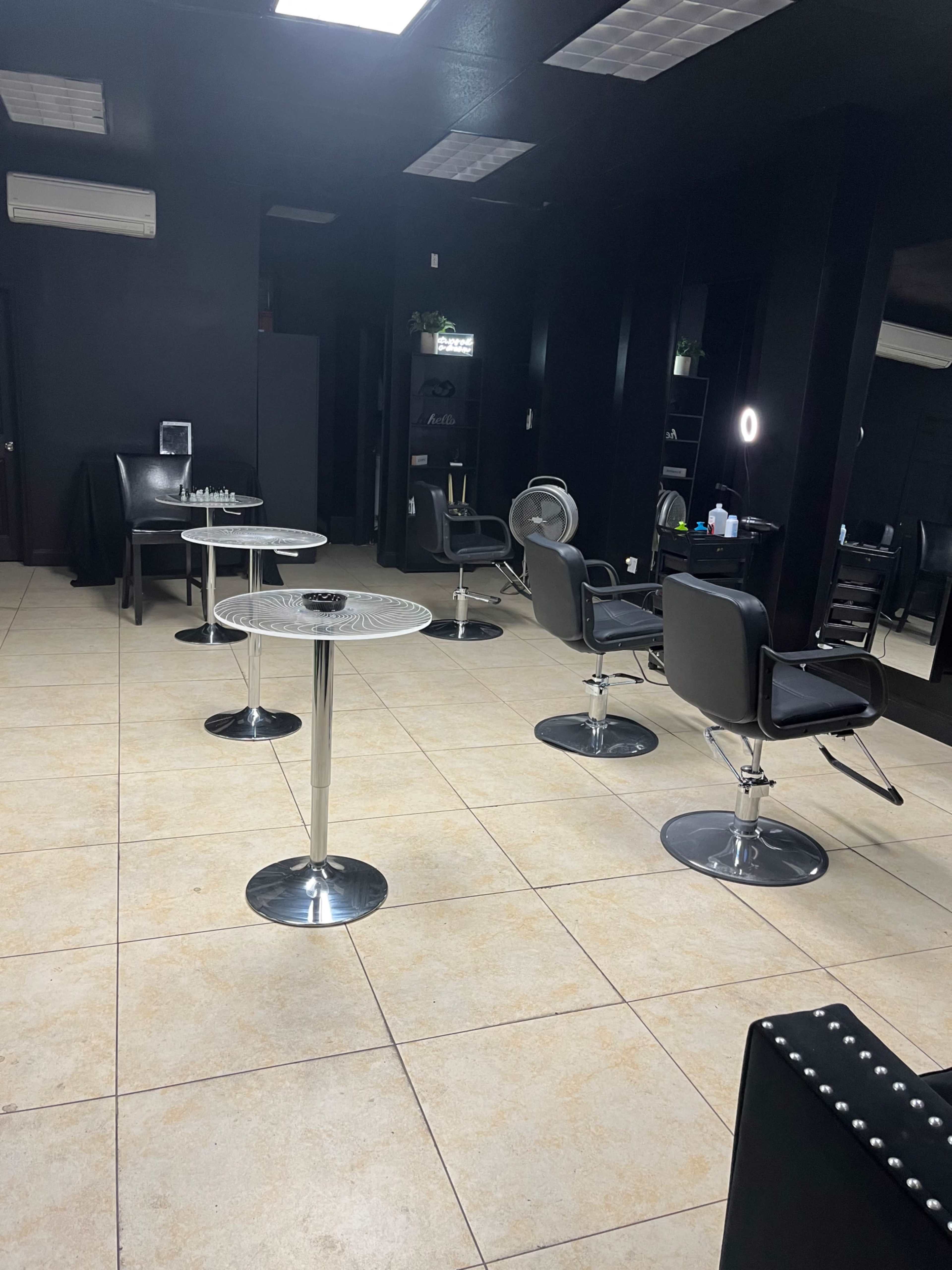 A hair salon features several black styling chairs and small tables arranged on a tiled floor in a darkly painted interior.