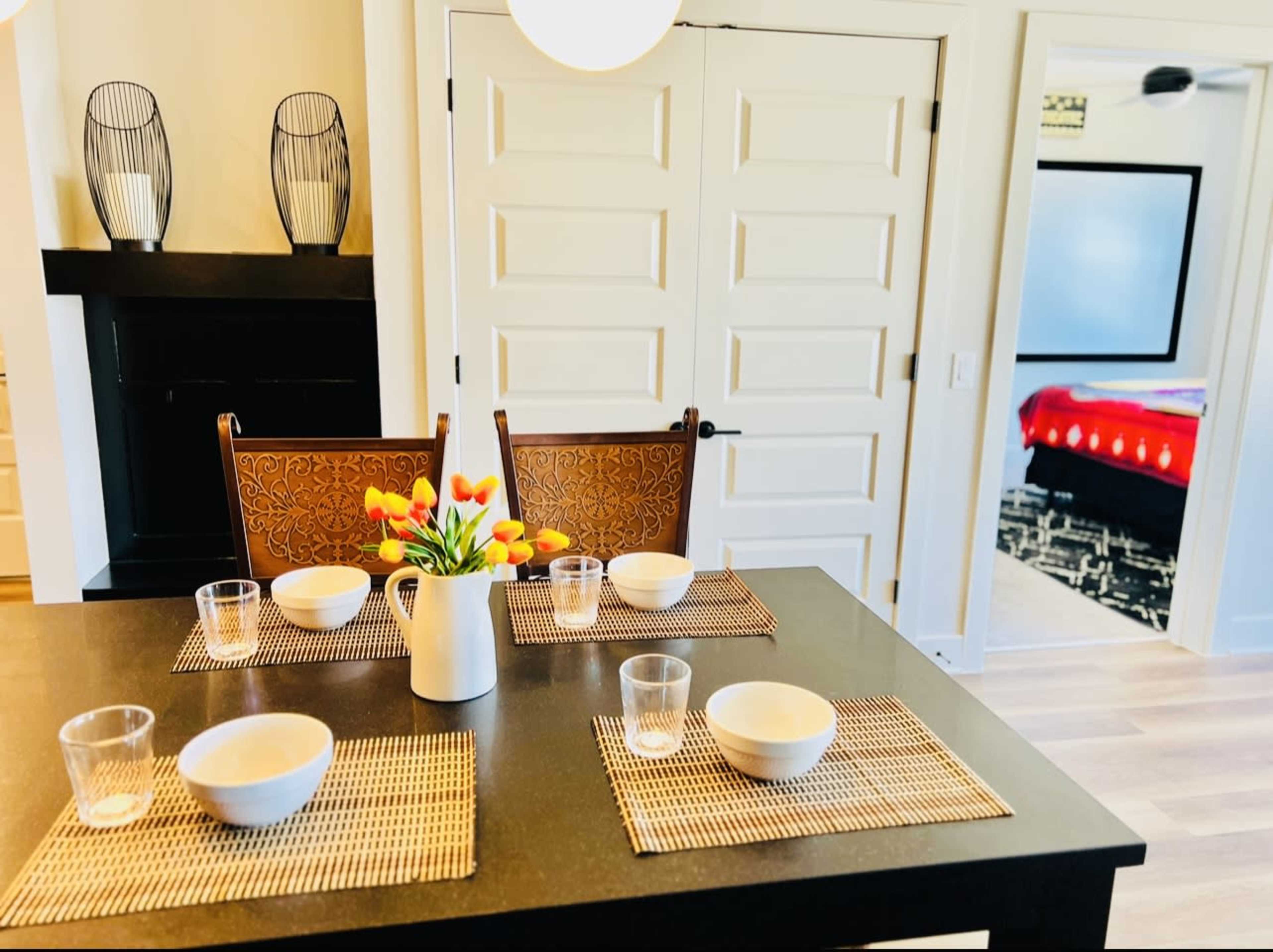 A dining table is set with four bowls and glasses, accompanied by a vase of flowers, in a room with light-colored walls and a glimpse of another room beyond.