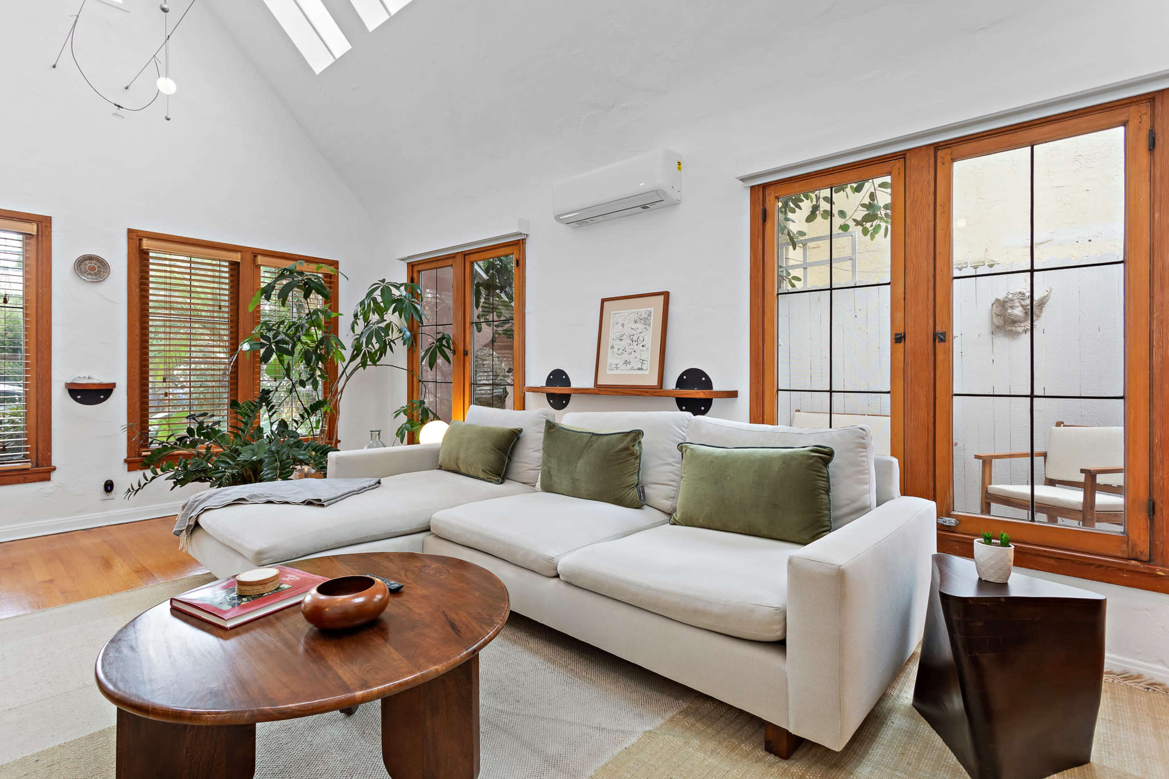 The image shows a bright living room with a white couch, wooden accents, large windows, and a small round wooden table.