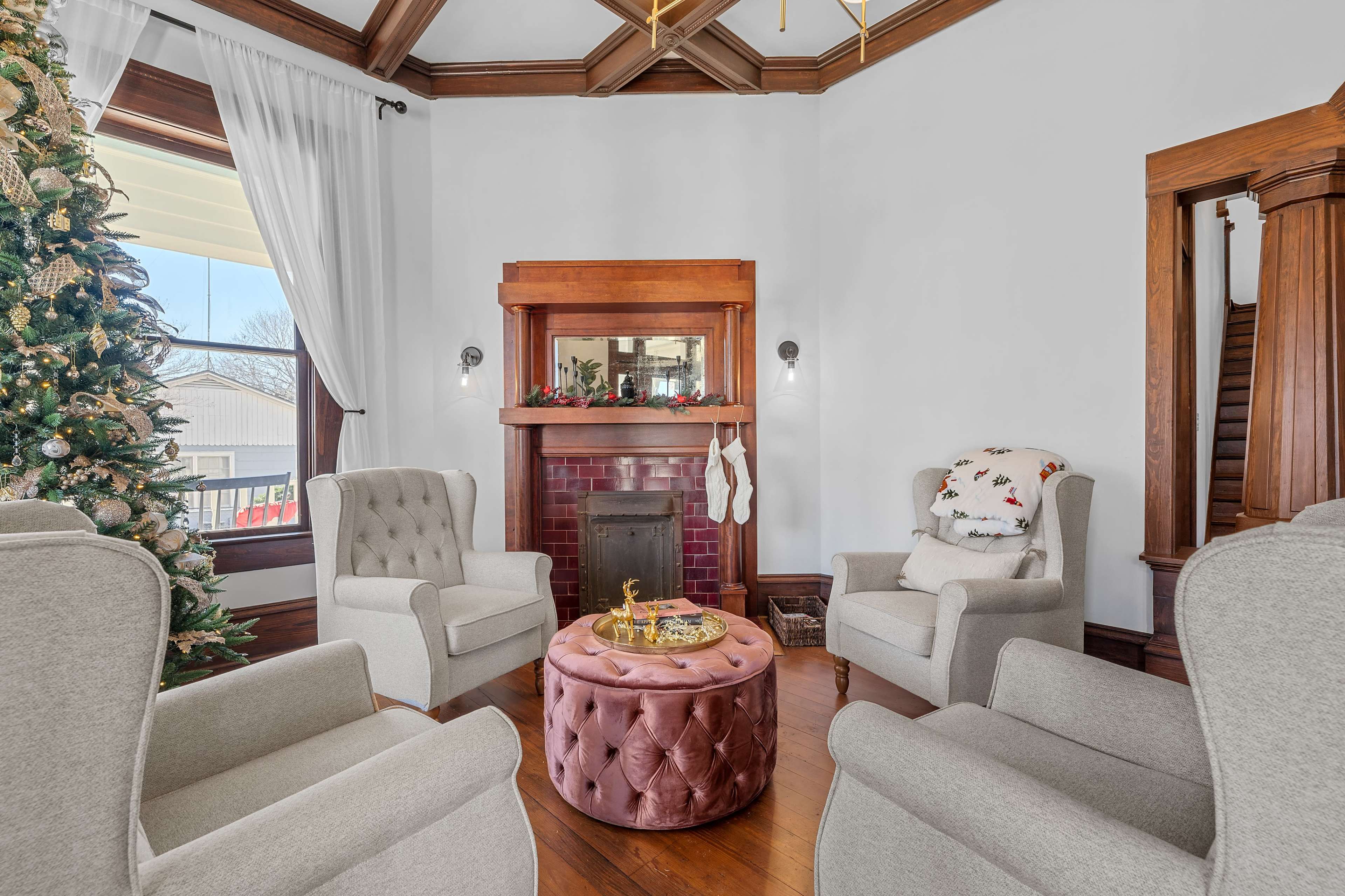 A well-furnished living room featuring a Christmas tree, a fireplace with decorative stockings, and four armchairs arranged around a round, tufted table.