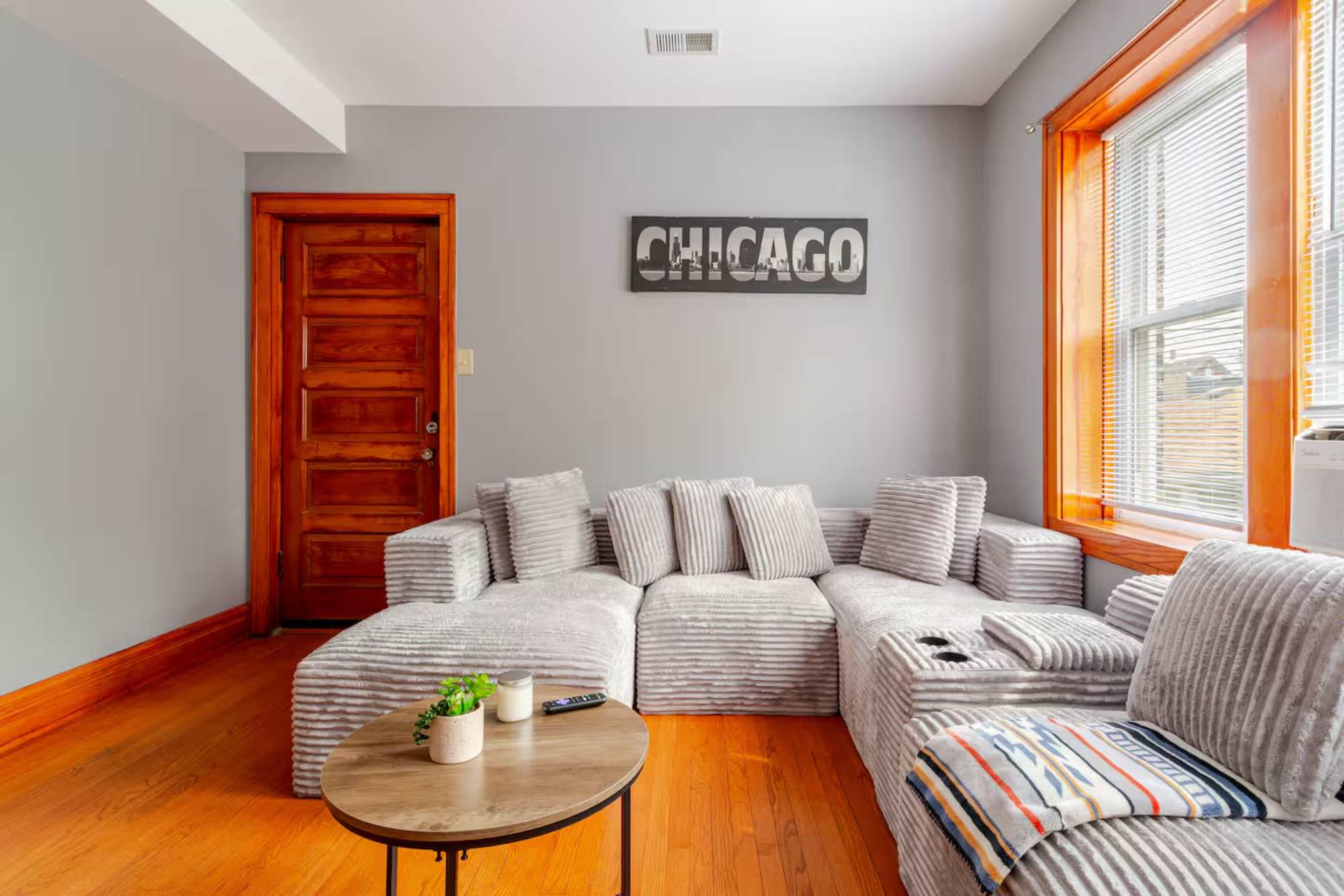 The image shows a cozy living room with a modern, striped sectional sofa, a round coffee table, a wooden door, and a "Chicago" wall art piece.