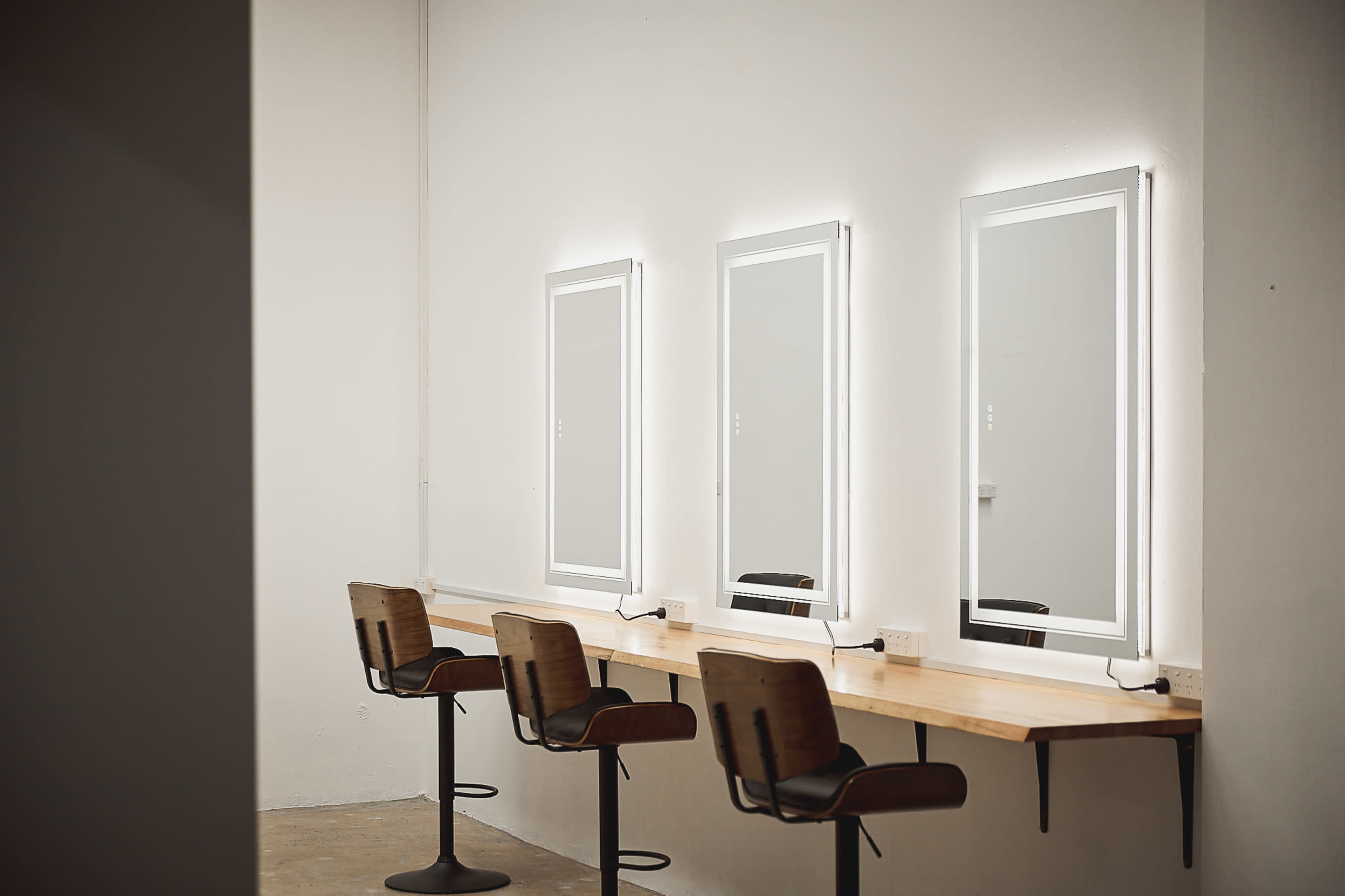 A minimalistic room with three illuminated mirrors above a wooden countertop and three tall stools.