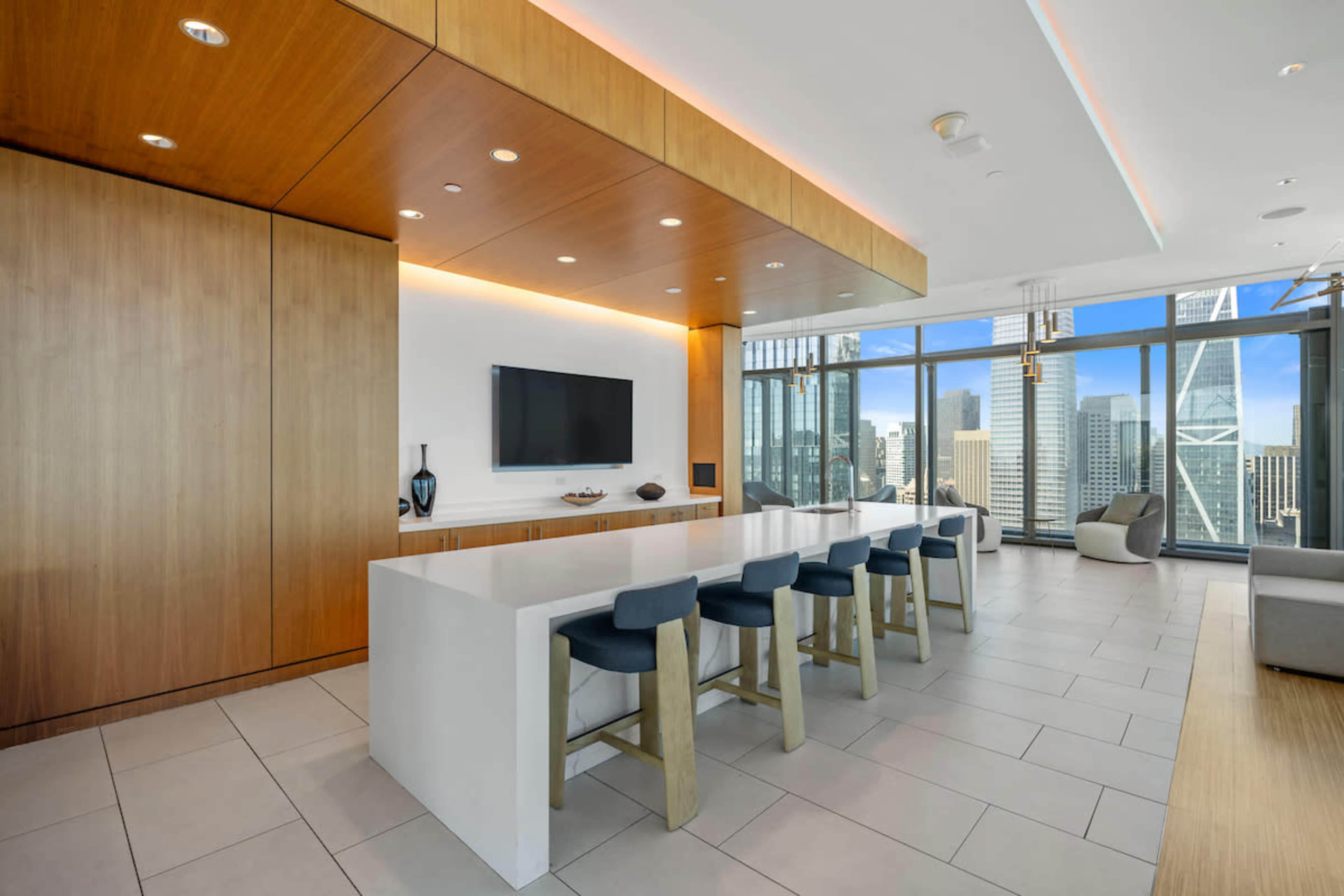 The image shows a modern kitchen area with a sleek white island and bar stools, large windows offering a view of a city skyline, and wooden accents on the walls and ceiling.