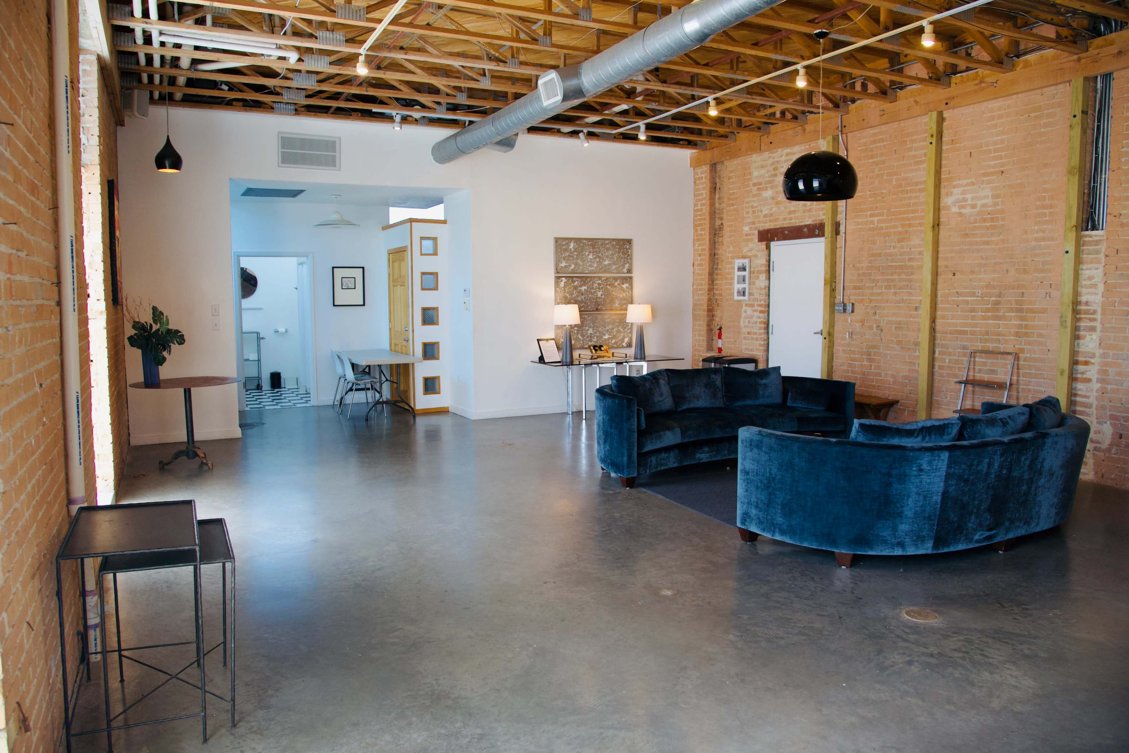 The spacious room features exposed brick walls, a concrete floor, and a modern circular blue sofa, with a dining area visible in the background.