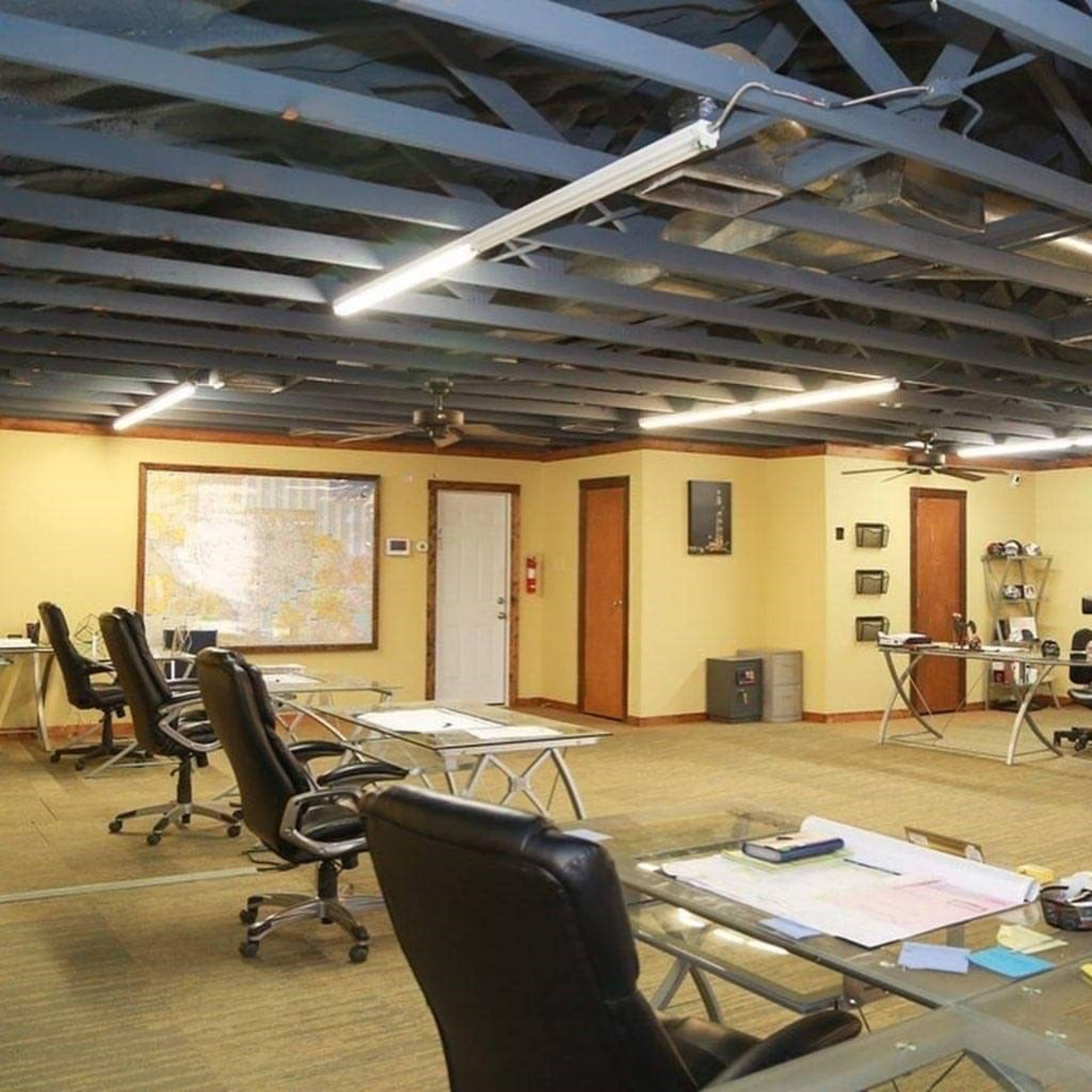 The image shows a spacious office with several glass-top desks and black chairs, featuring a map on the wall and an open ceiling with exposed beams.