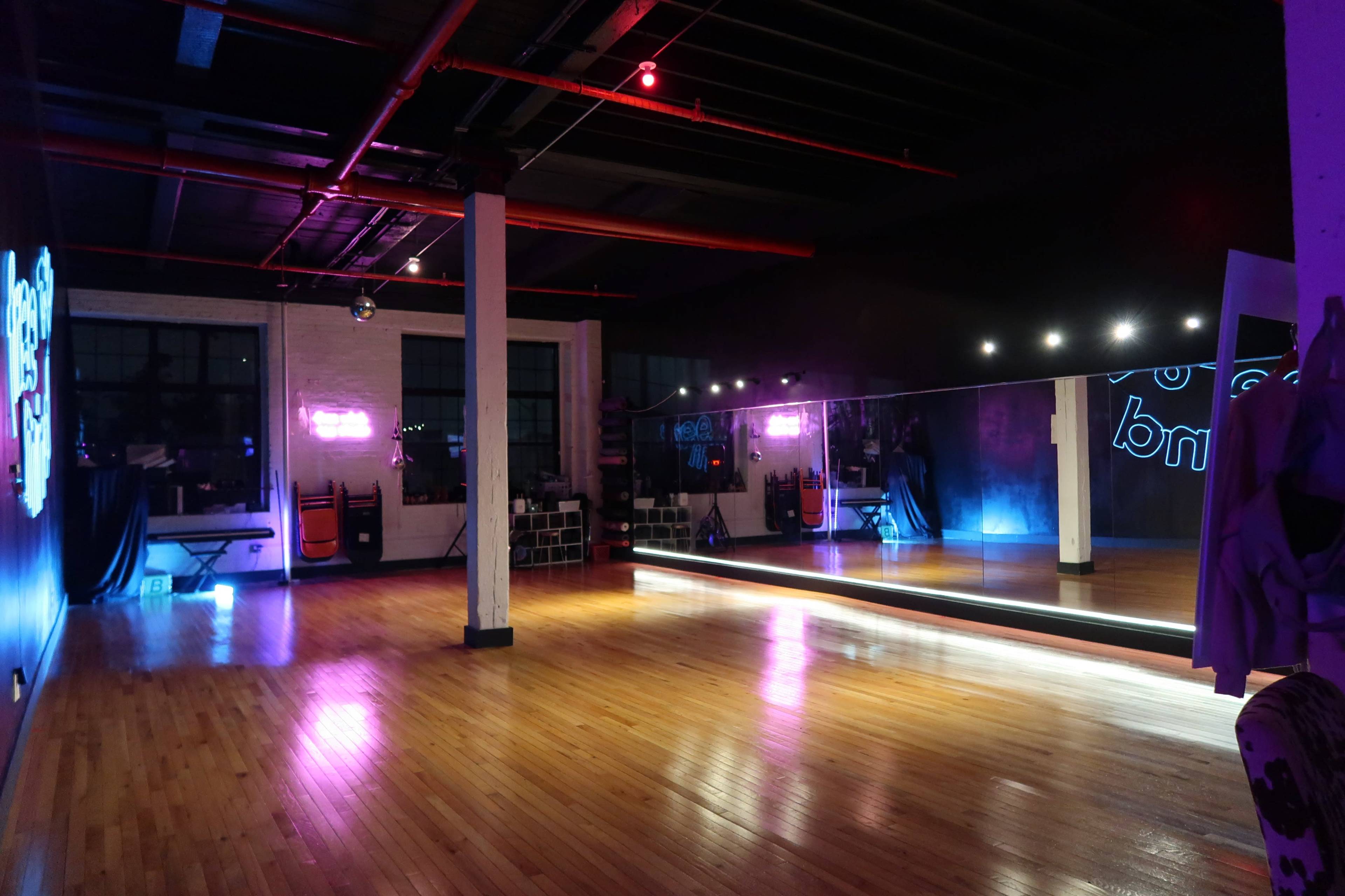 The image shows an empty dance studio with wooden floors, a large mirror, and neon lights illuminating the space.