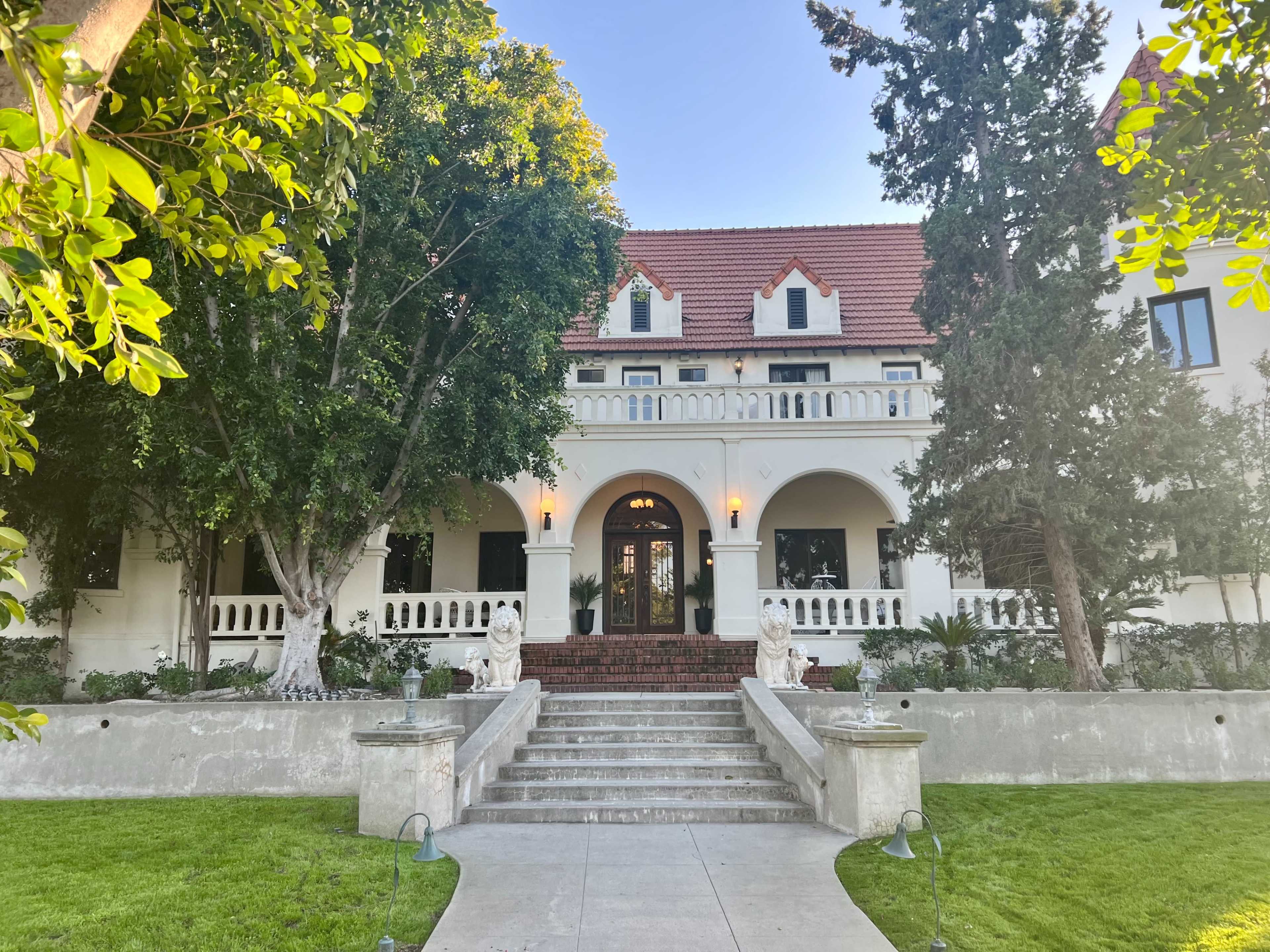 A large, elegant house with a red-tiled roof, flanked by trees, and featuring a wide staircase leading to a front entrance with arched doorways.