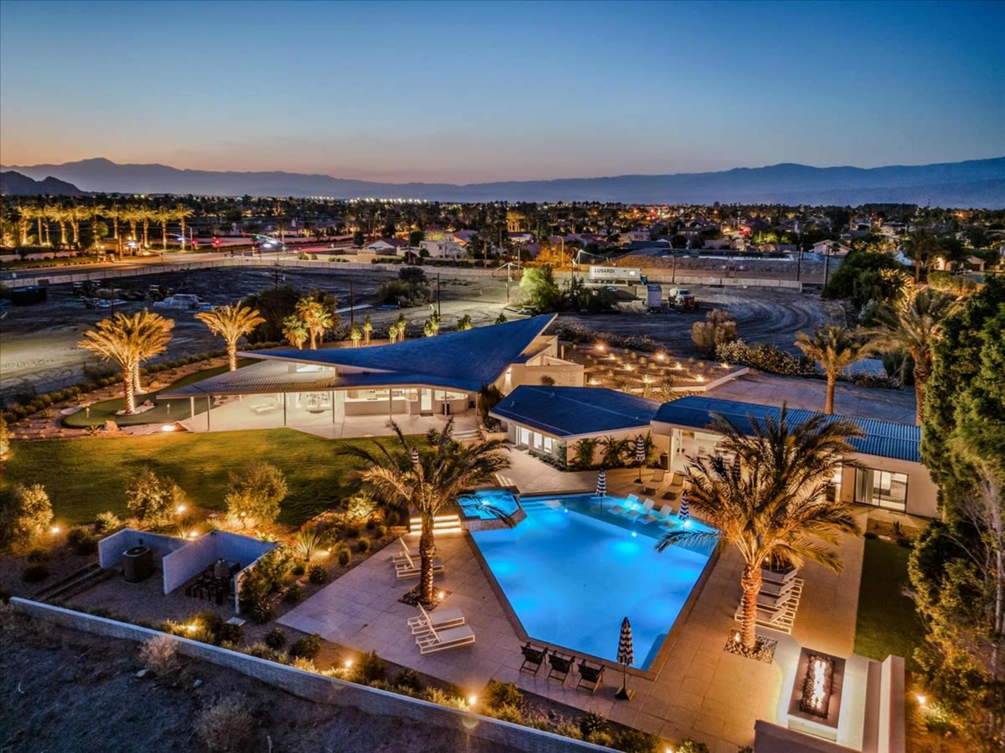 A modern home with a large swimming pool and palm trees is illuminated at dusk, surrounded by open land and distant mountains.