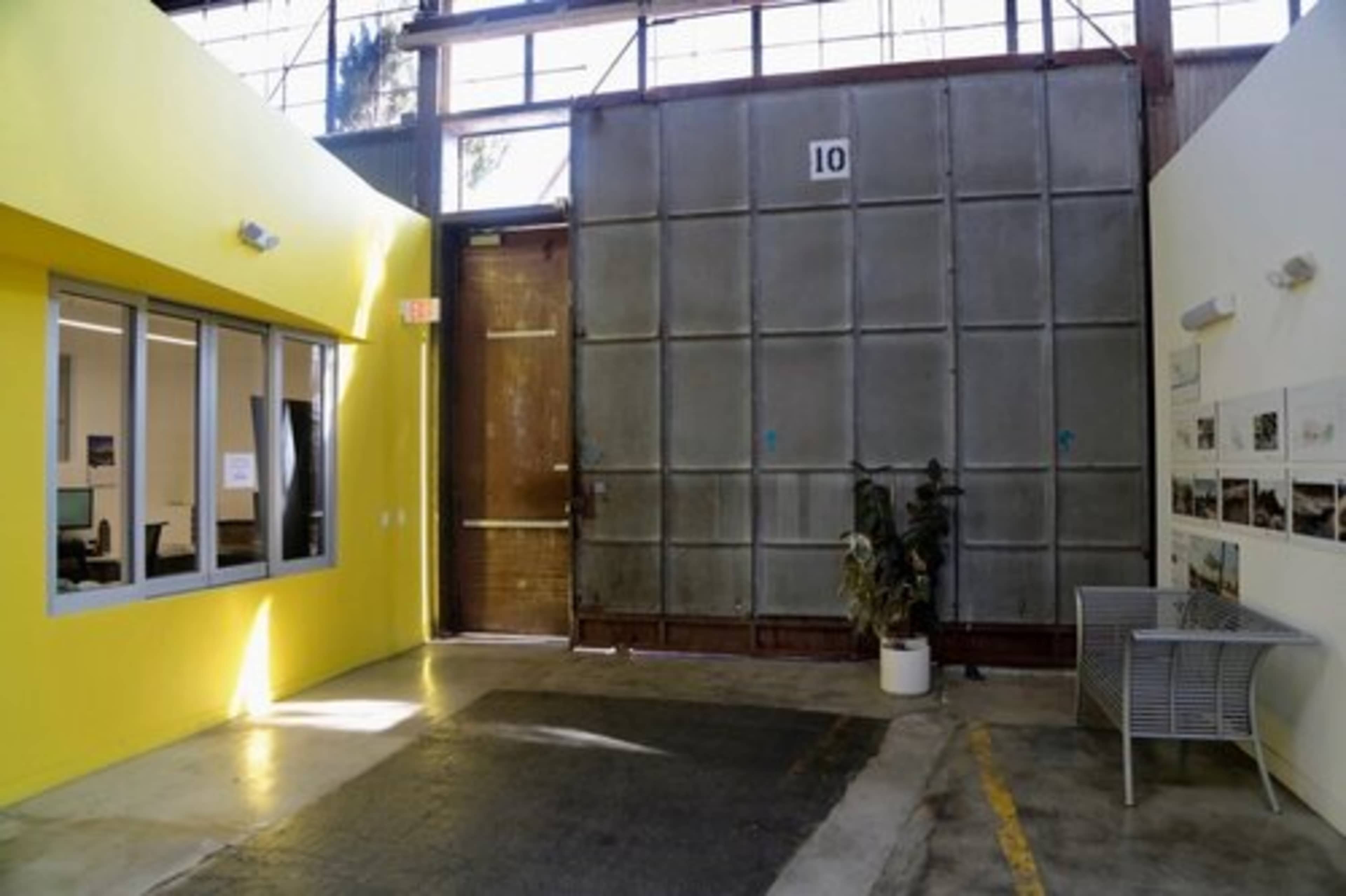 The image shows an industrial interior with a large metal door, a yellow wall, a windowed office space, and a potted plant near a bench.