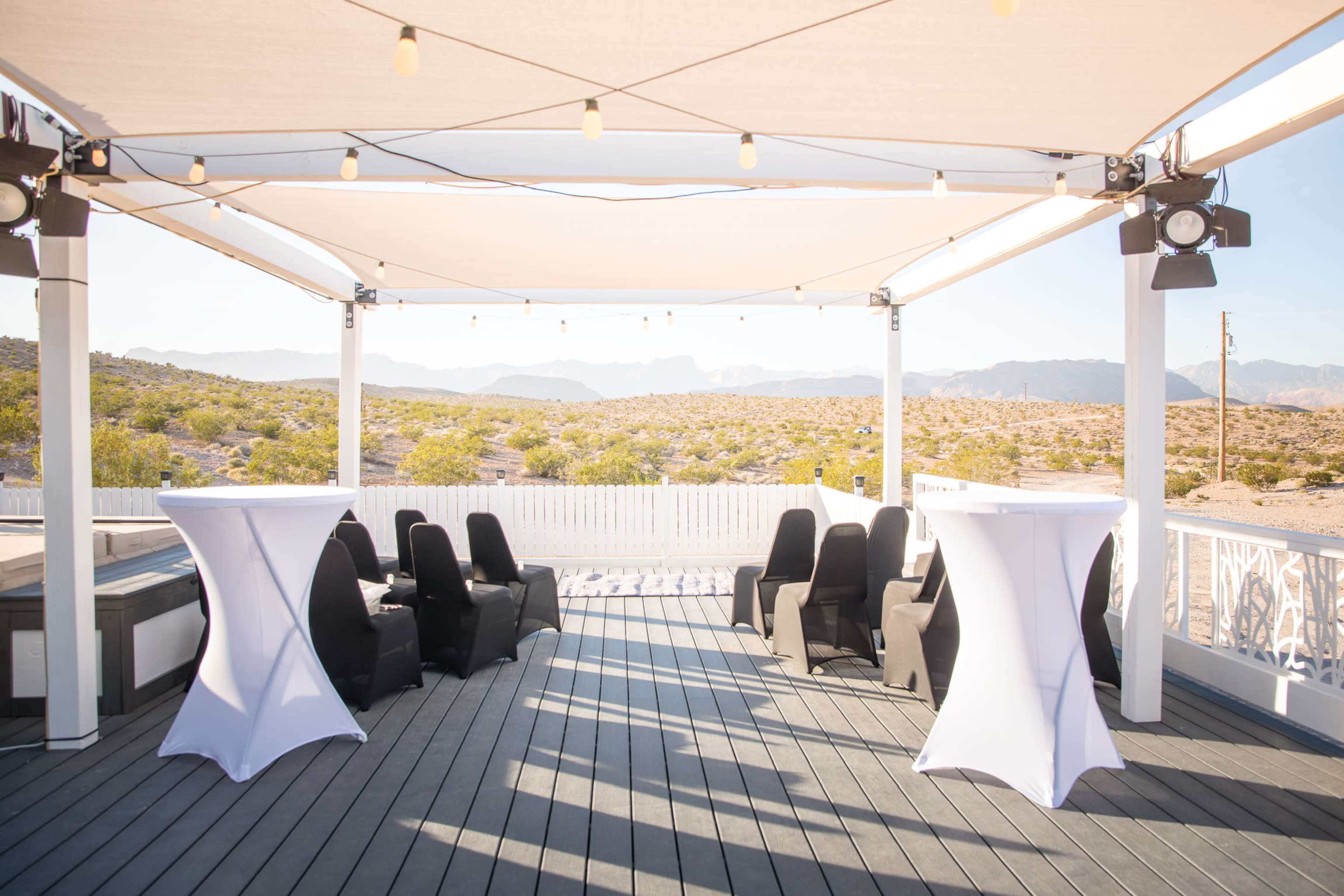 A terrace area features black chairs and white cocktail tables under a shaded canopy with string lights, overlooking a desert landscape.