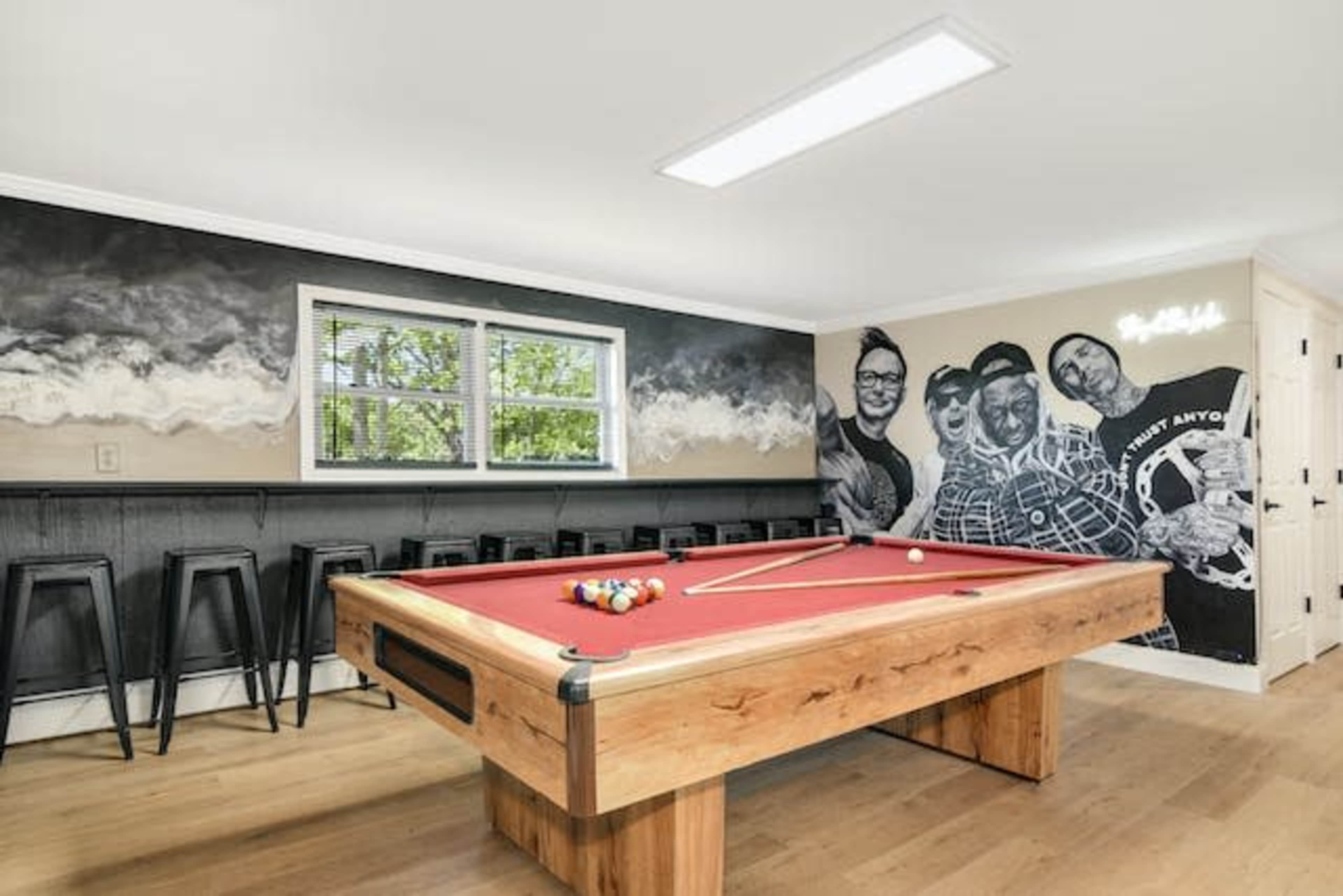 The image shows a game room featuring a red pool table with a wooden frame, surrounded by black bar stools and a mural of three figures on the wall.