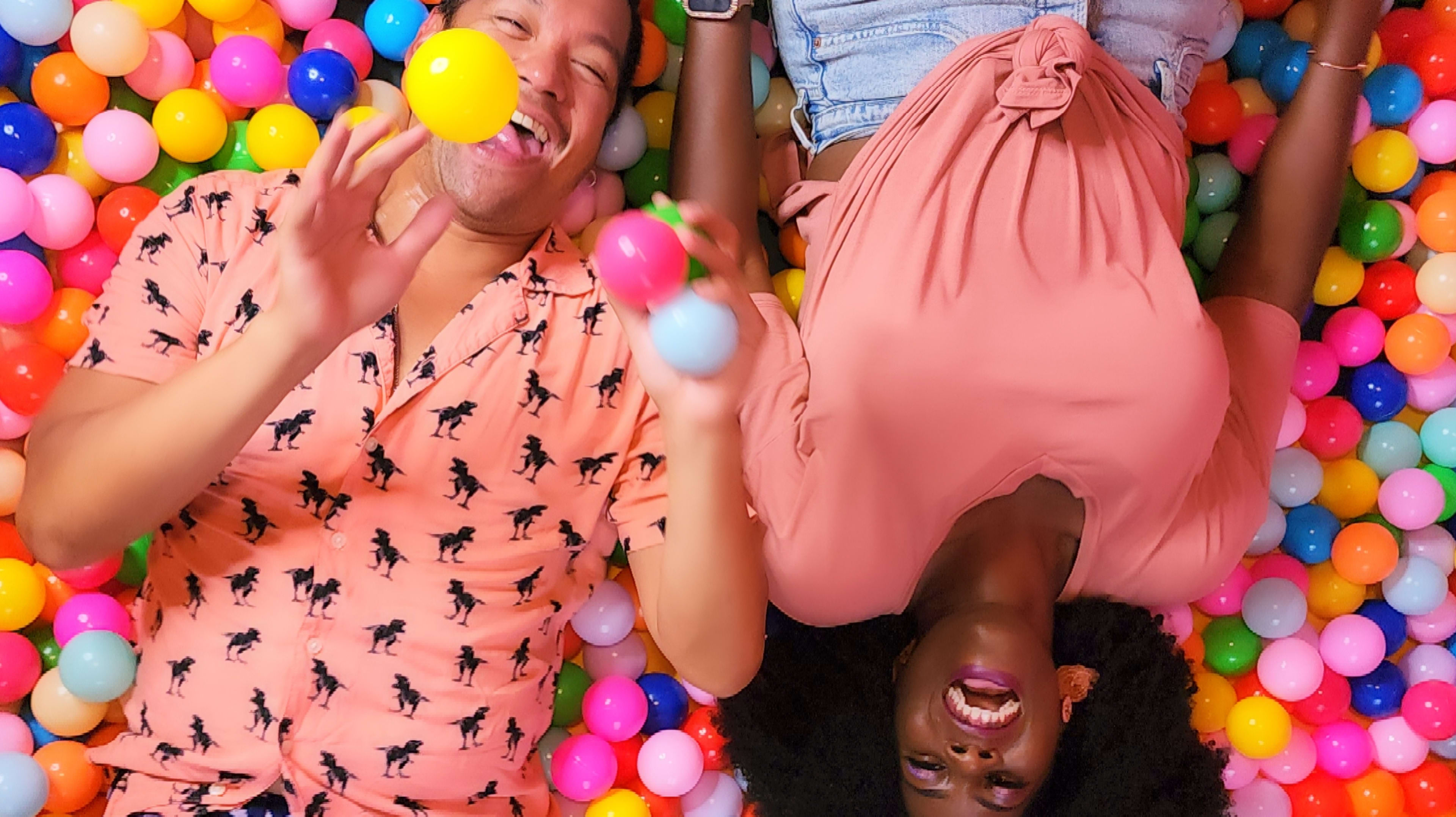 Two people lie on a colorful floor covered with plastic balls, laughing while holding balls in their hands.