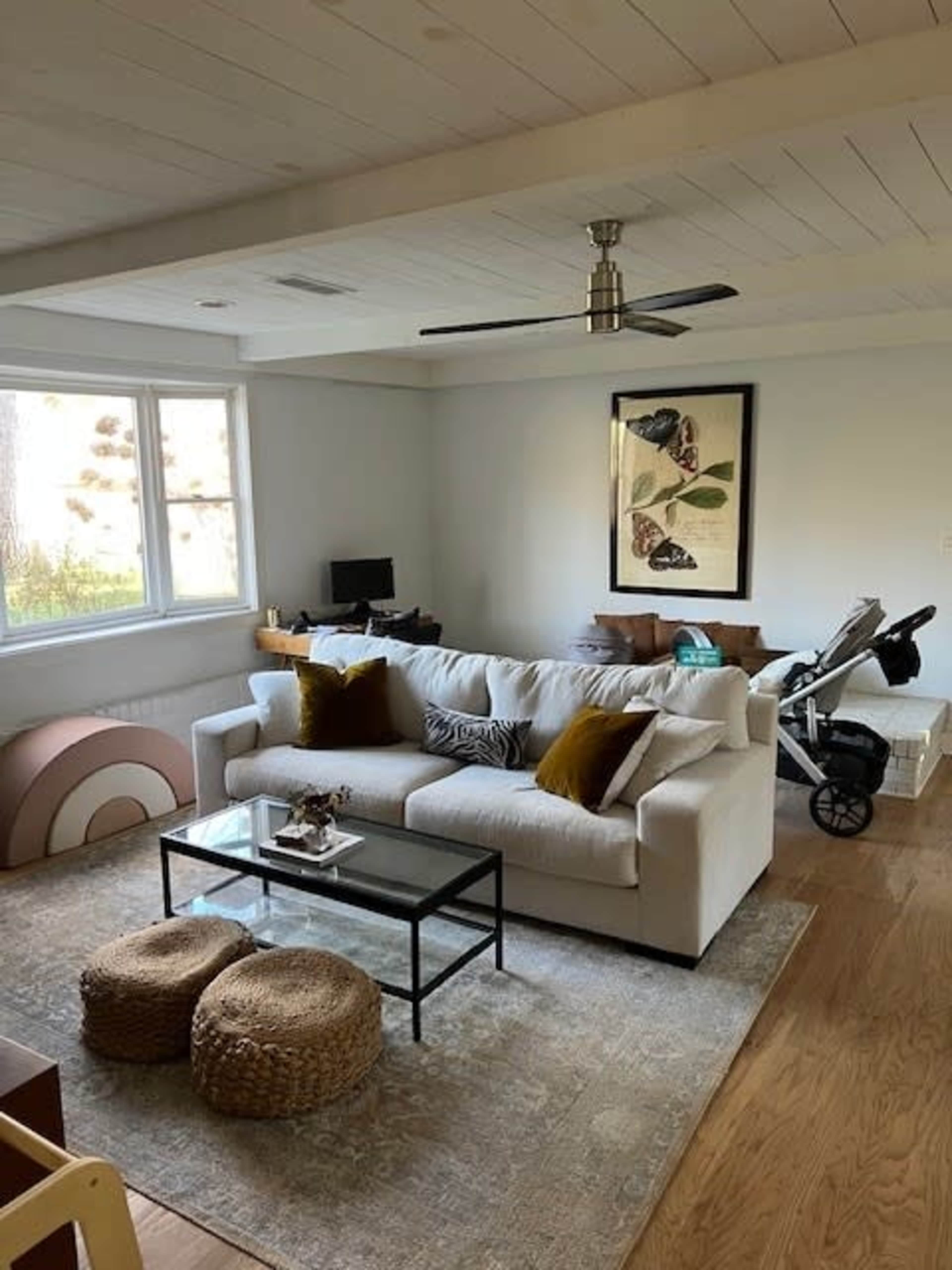A modern living room with a white sofa, a glass coffee table, and a large window, featuring a blend of neutral decor and a baby stroller in the corner.