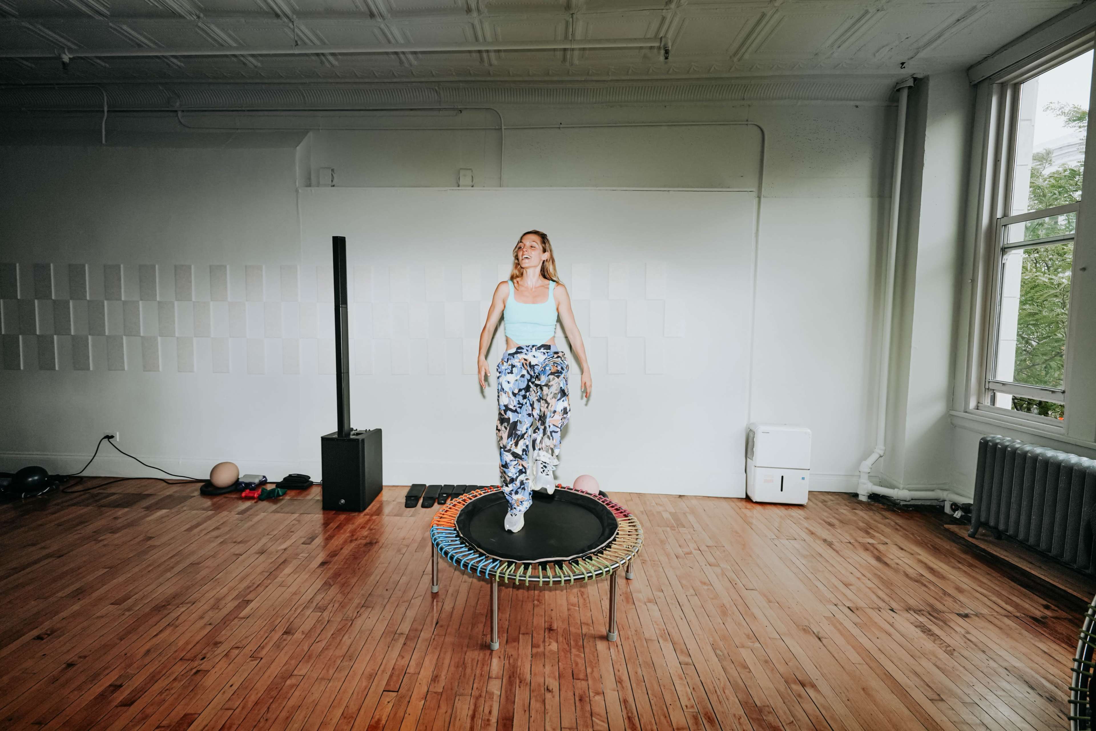 A person jumps on a trampoline in a spacious room with wooden floors and large windows.