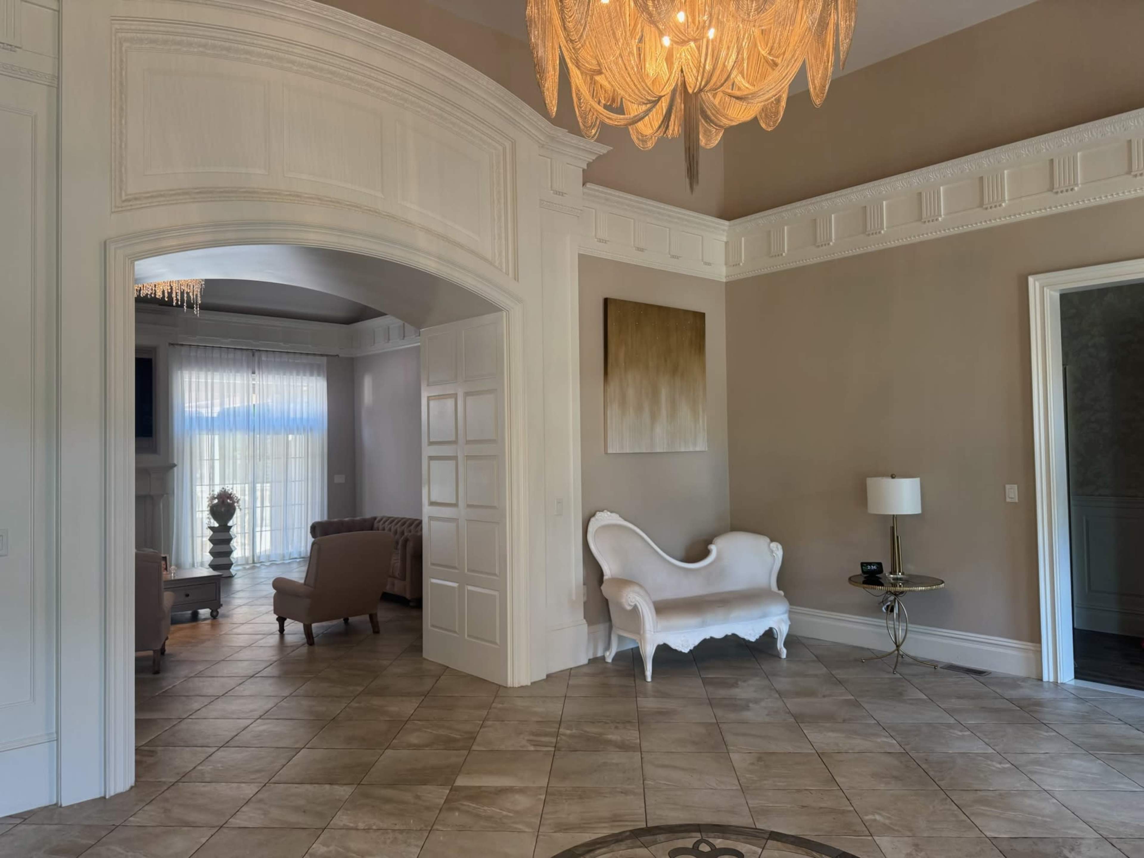 The image shows an elegantly designed foyer with a chandelier, a white seating area, and adjacent rooms featuring large windows and neutral-colored walls.