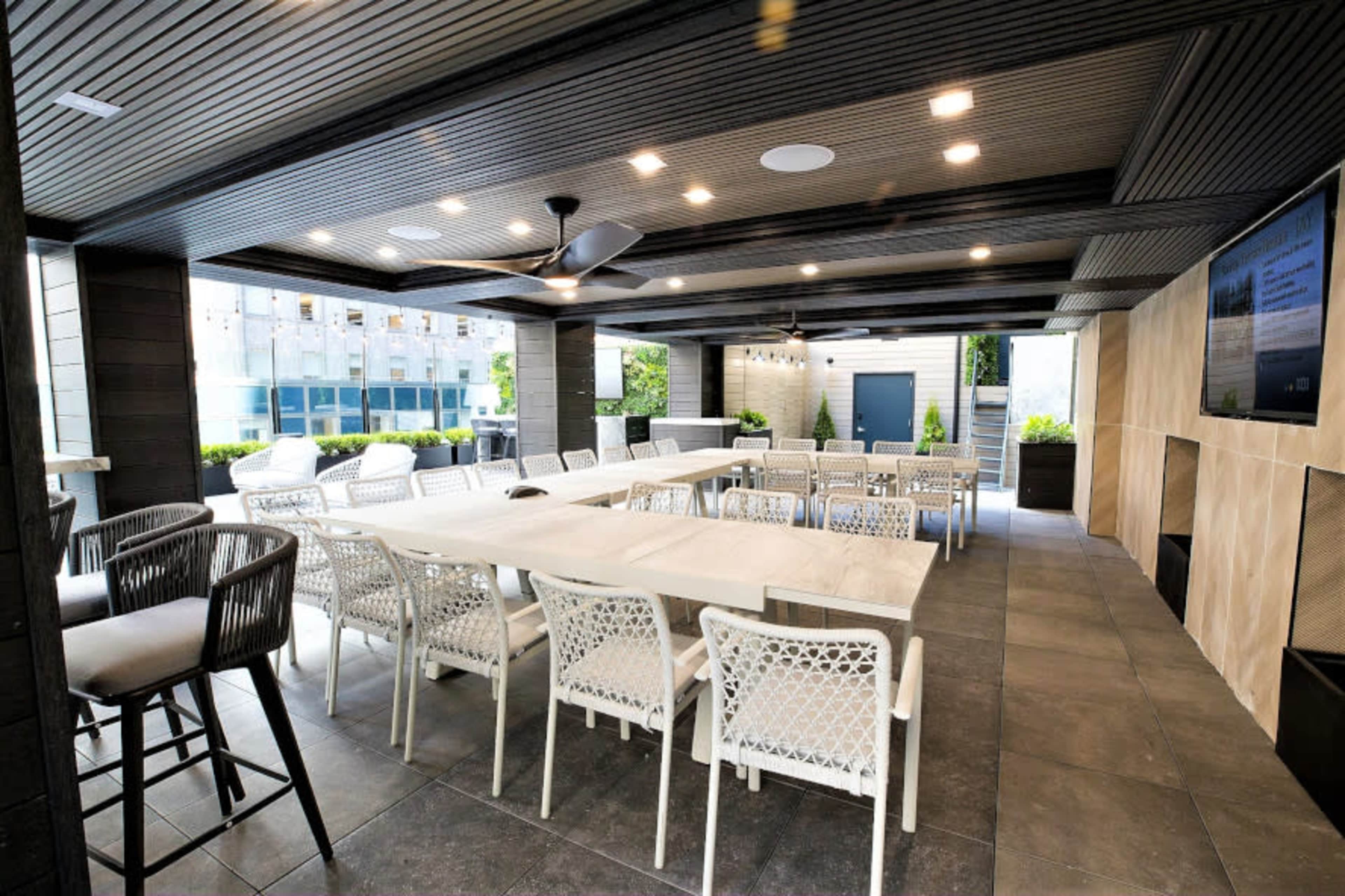 The image shows a modern outdoor dining area with a large table surrounded by white chairs, a ceiling fan, and large windows letting in natural light.