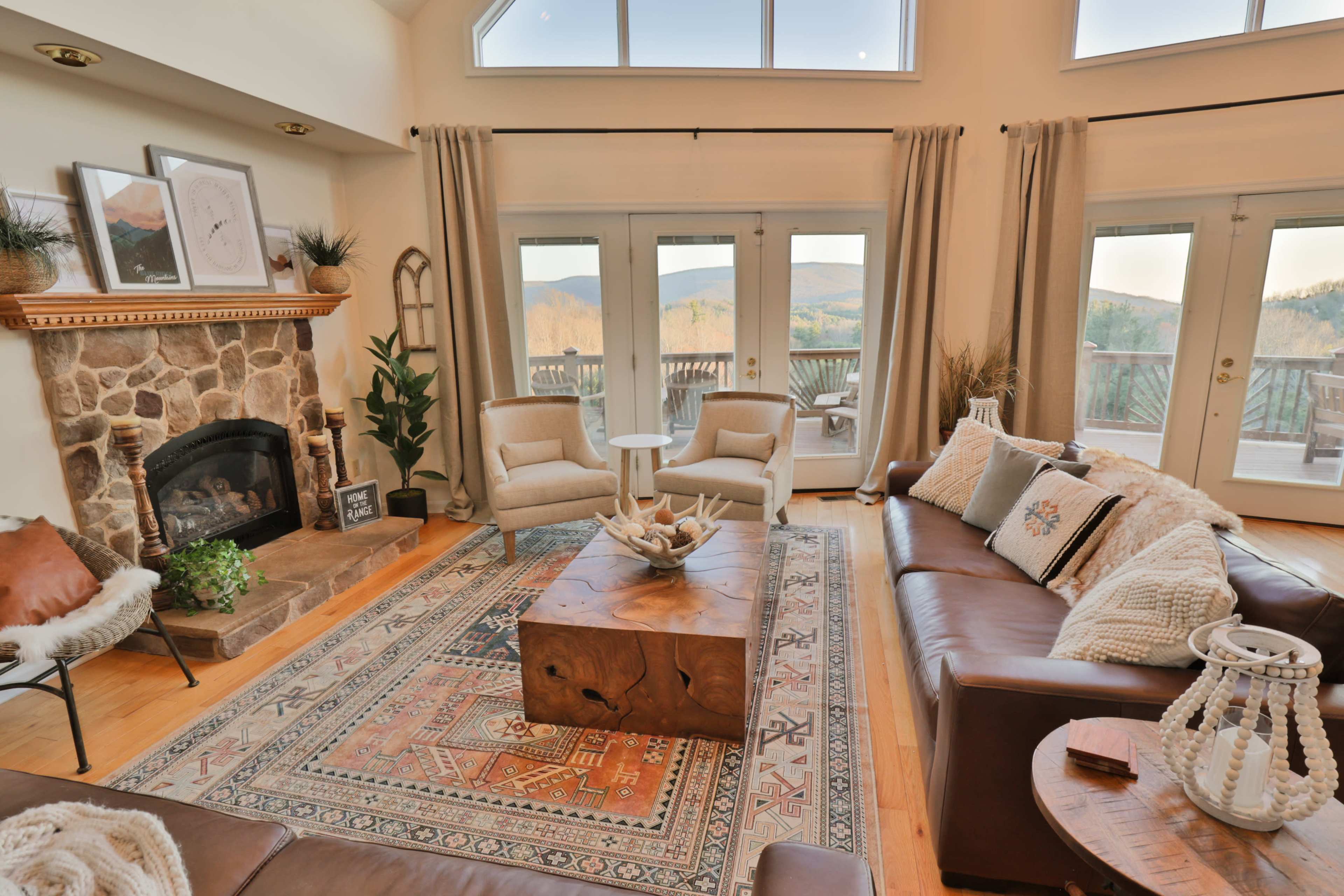 The living room features a stone fireplace, a wooden coffee table, two armchairs, and large windows overlooking a mountainous landscape.
