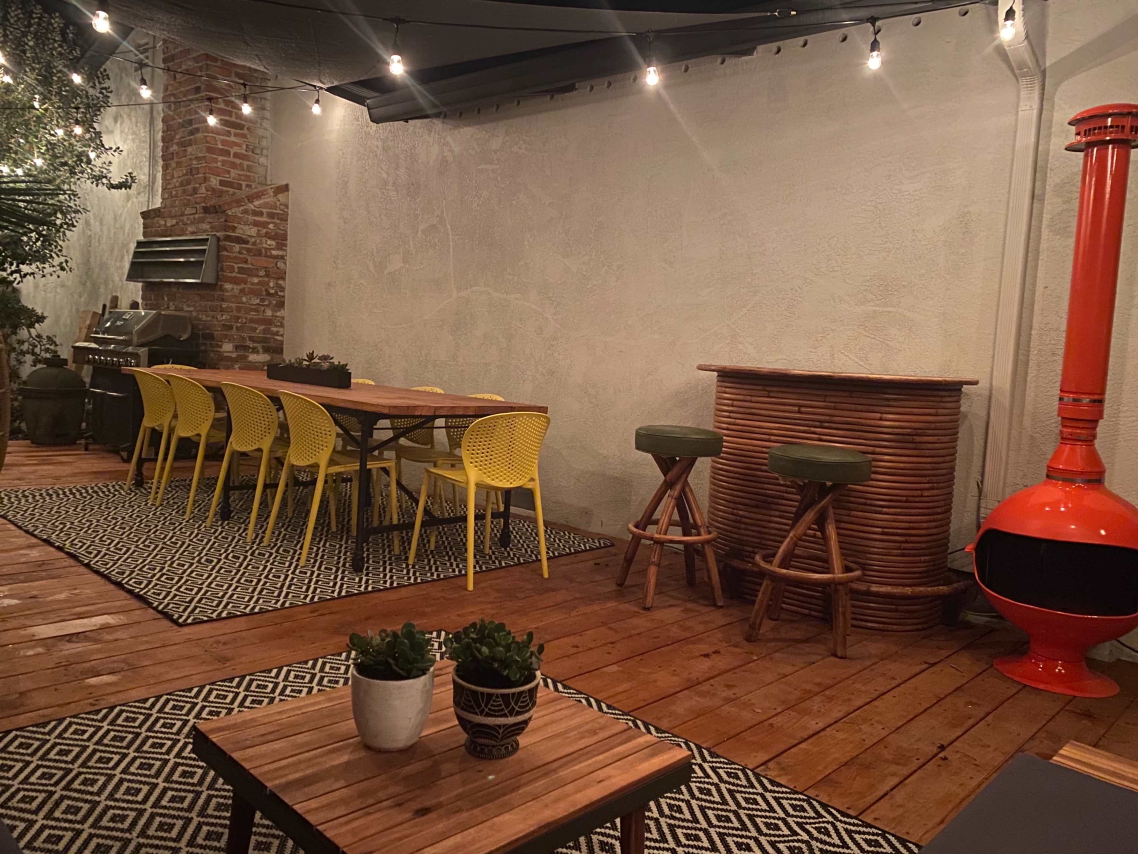 A patio area featuring a long dining table with yellow chairs, a grill, a bar with stools, and decorative plants, all under ambient lights.