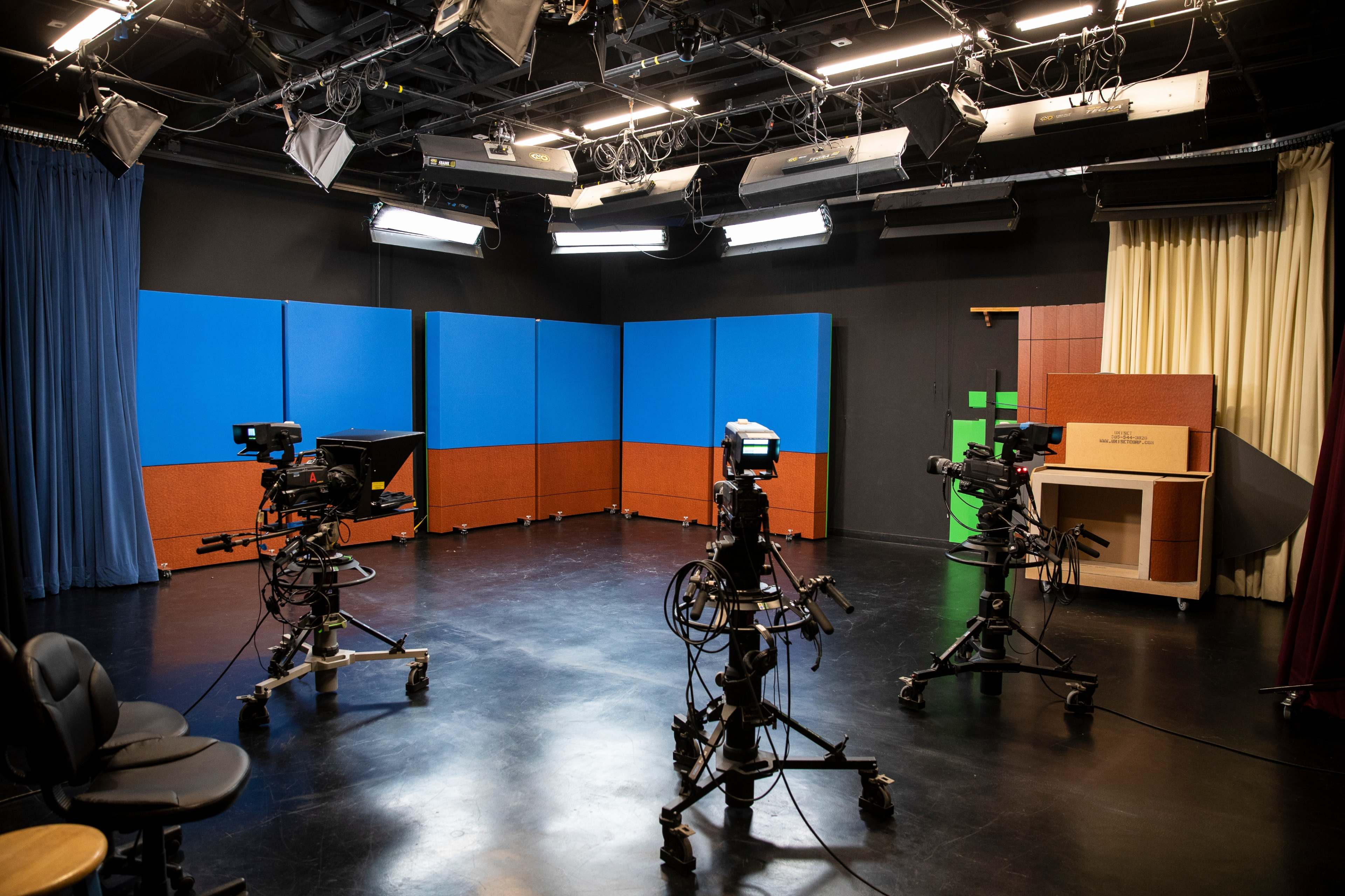 A television studio is set up with multiple cameras facing a blue backdrop and equipment, surrounded by studio lights and seating.